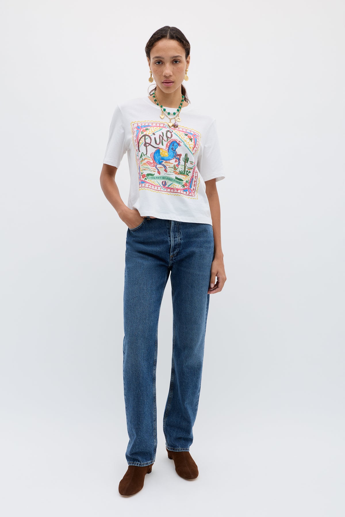 A woman stands against a plain white background, wearing the Ria - Rider White T-shirt with colorful artwork, straight blue jeans, brown shoes, and a beaded necklace. She has one hand in her pocket and a neutral expression.