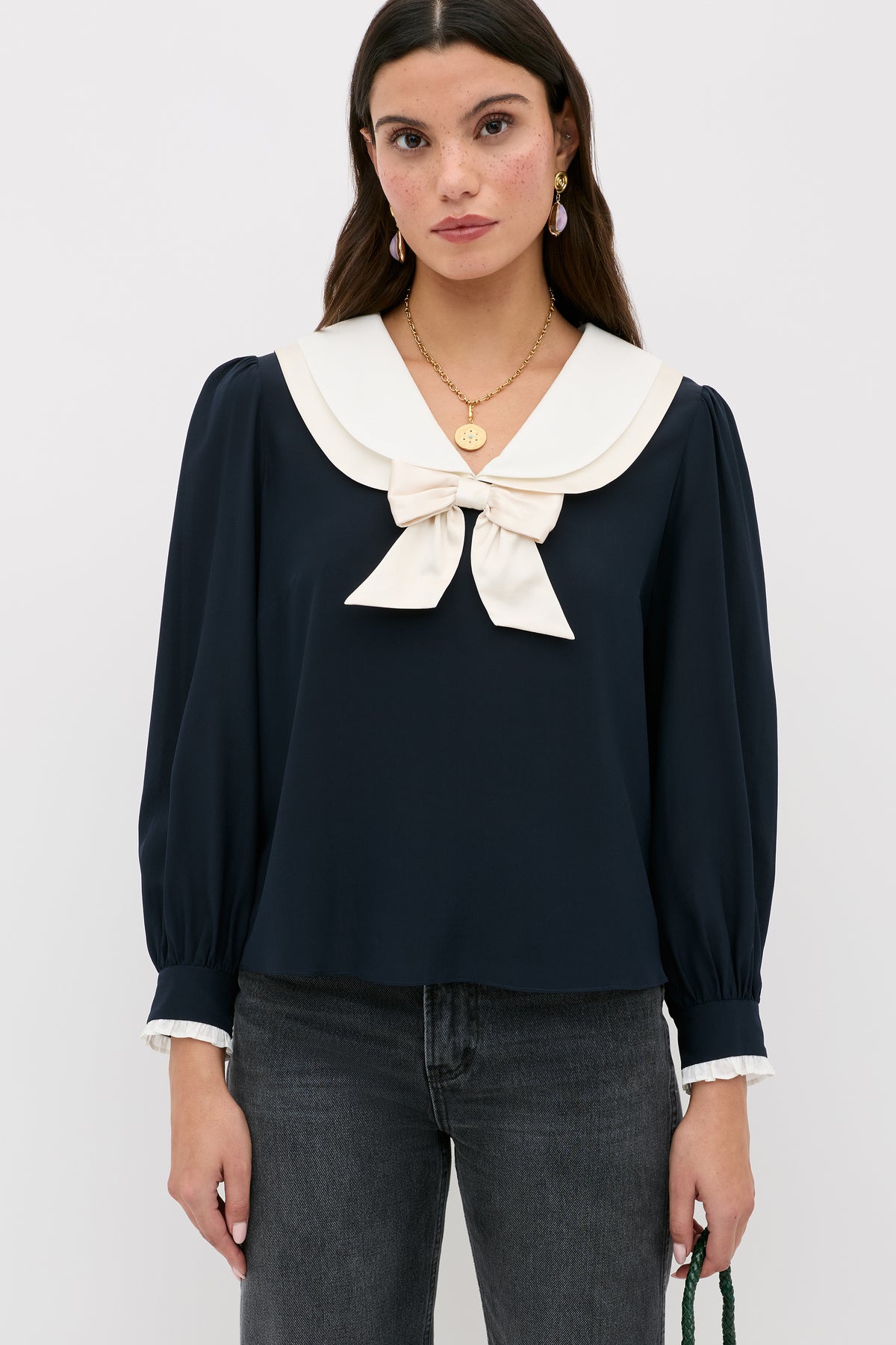 A woman with long brown hair wears the Eimer - Navy silk top featuring a large sailor collar, bow, blouson sleeves, and dark jeans. She stands against a white background and accessorizes with gold jewelry.