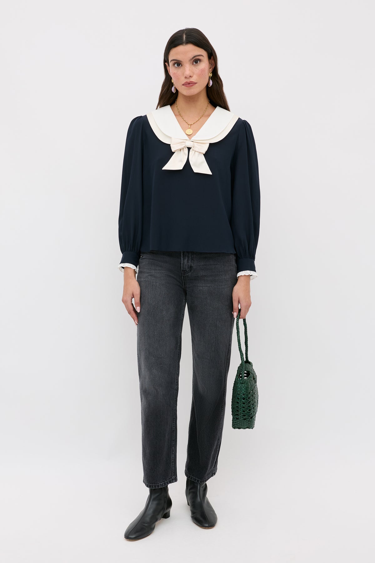 A woman stands against a white background wearing the Eimer - Navy blouse with a sailor-style collar and bow, dark gray jeans, black ankle boots, and holding a green woven bag.