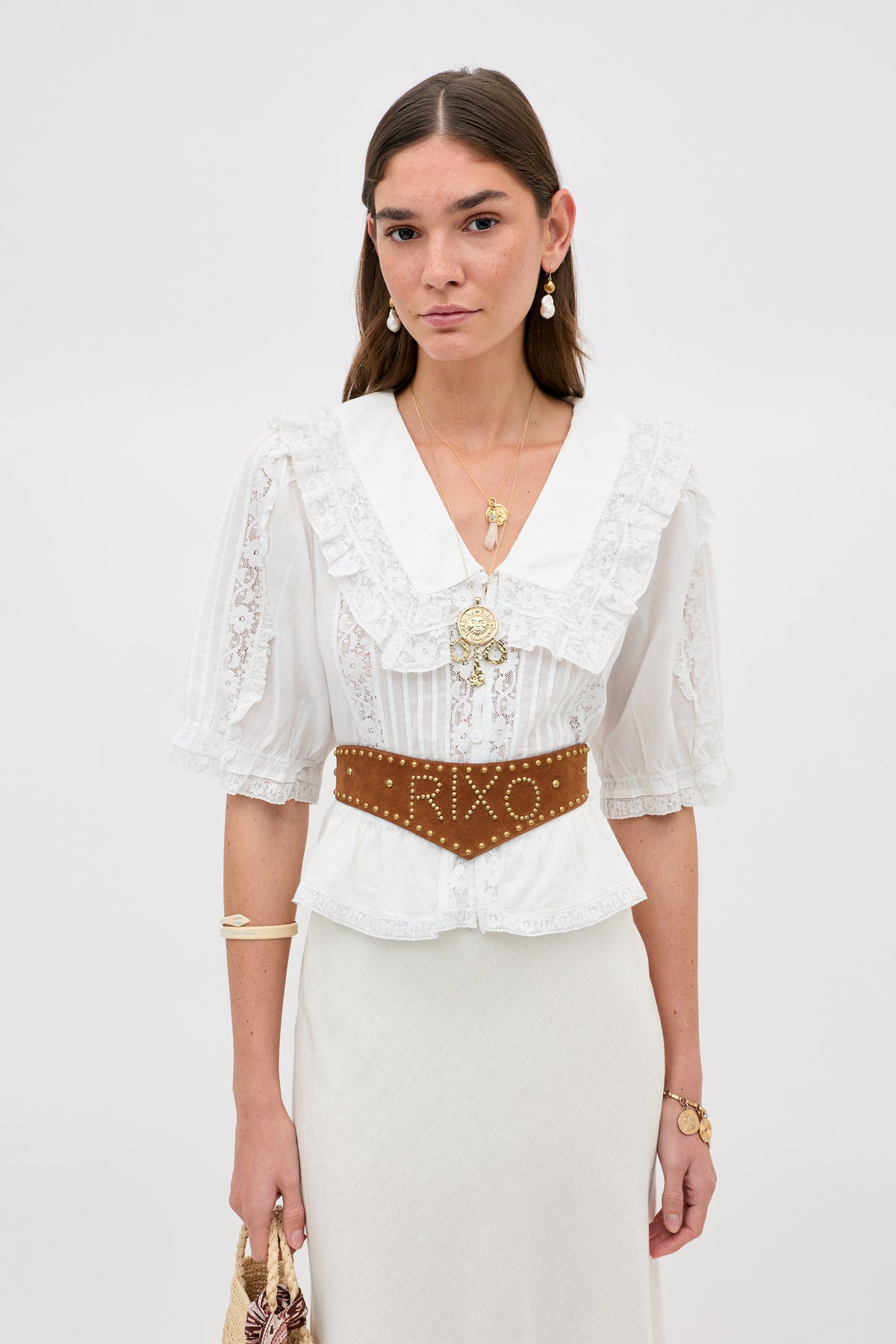A woman wears the Ellery - White vintage-inspired blouse with lace details and a ruffled collar, styled with layered gold jewelry and a brown belt. She carries a small handbag and stands against a plain background.