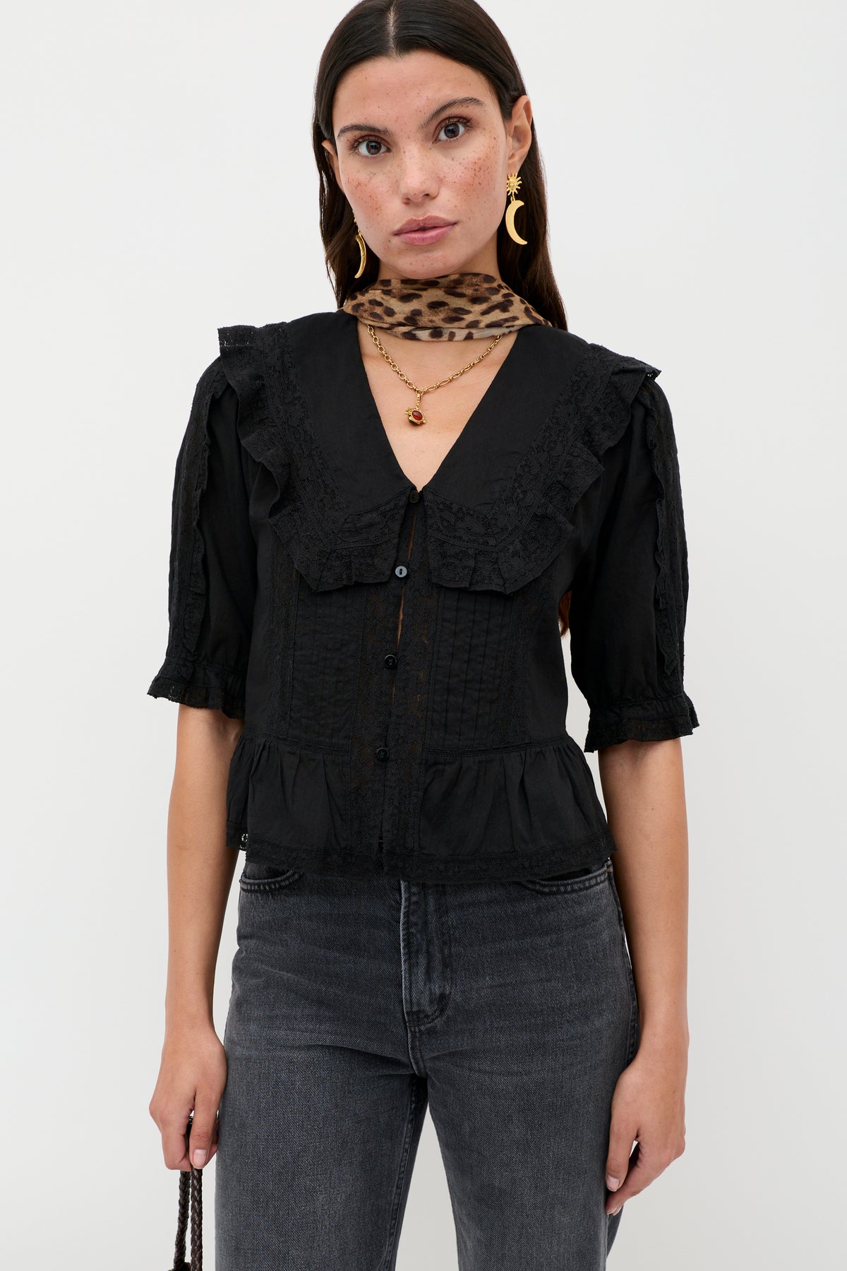 A woman in the Ellery - Black vintage-inspired cotton button-down with a ruffled collar, paired with dark gray jeans, gold jewelry, and a leopard print scarf, stands against a plain white background, looking at the camera.