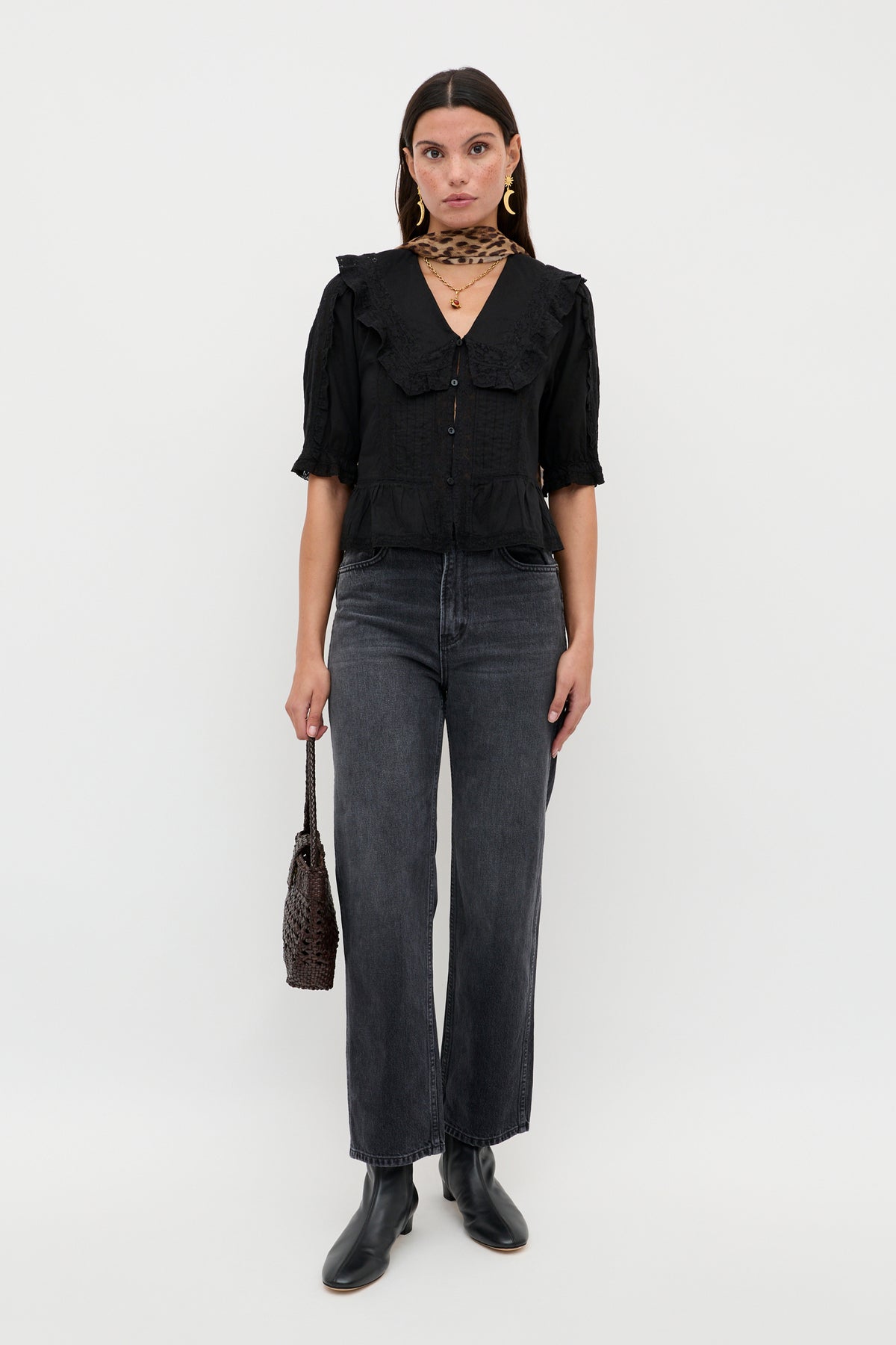 A woman stands facing forward against a plain white background, wearing the Ellery - Black vintage-inspired cotton button-down top with a wide collar, styled with dark gray jeans, black ankle boots, gold earrings, layered necklaces, and a woven handbag.