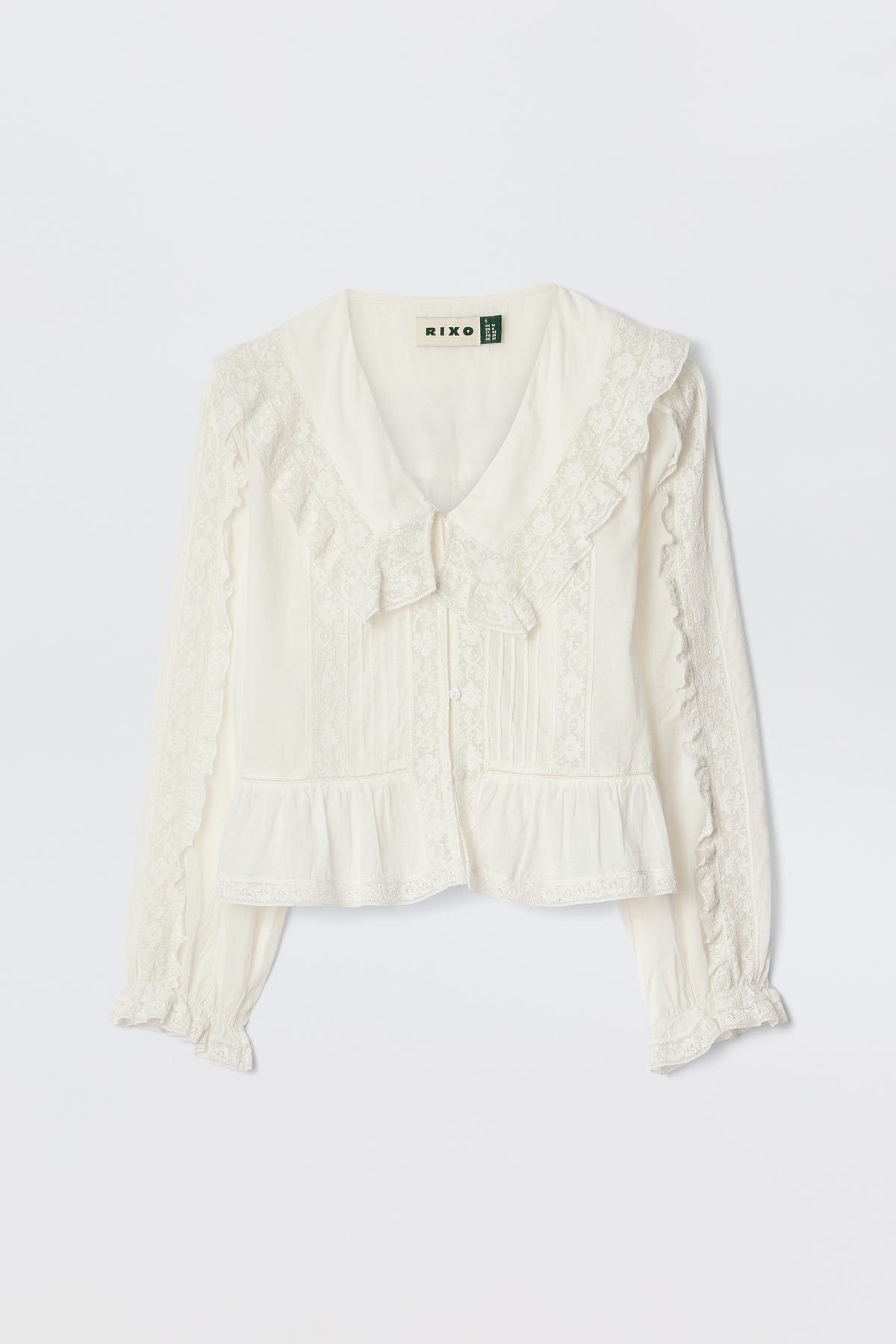 The Maribelle - Ivory is a vintage-inspired, long-sleeved white blouse with lace trim, button-down front, ruffled cuffs, and a wide collar with bow detail, shown on a white background.