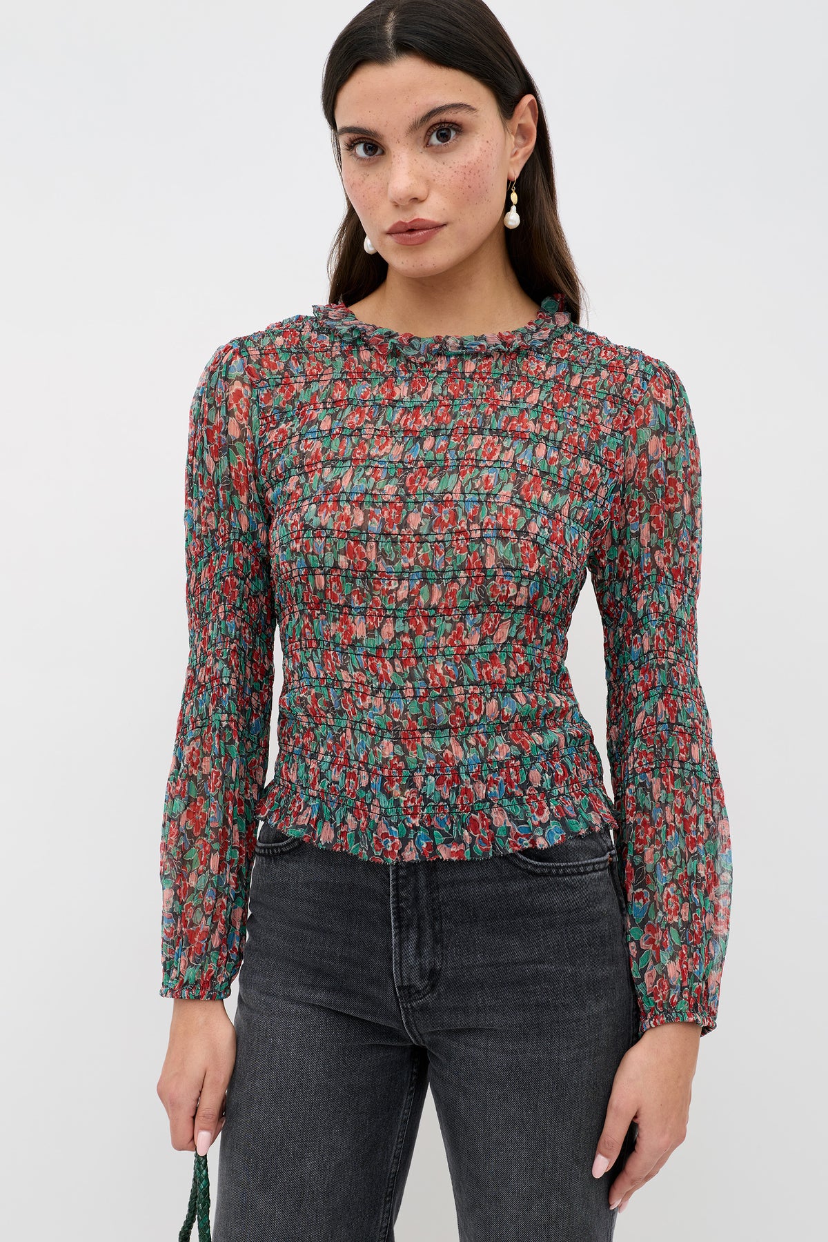 A woman wears the Gardenia - Jeannie Bloom Charcoal sheer, long-sleeve floral blouse with black jeans and drop earrings, standing against a white background and looking at the camera with a neutral expression.