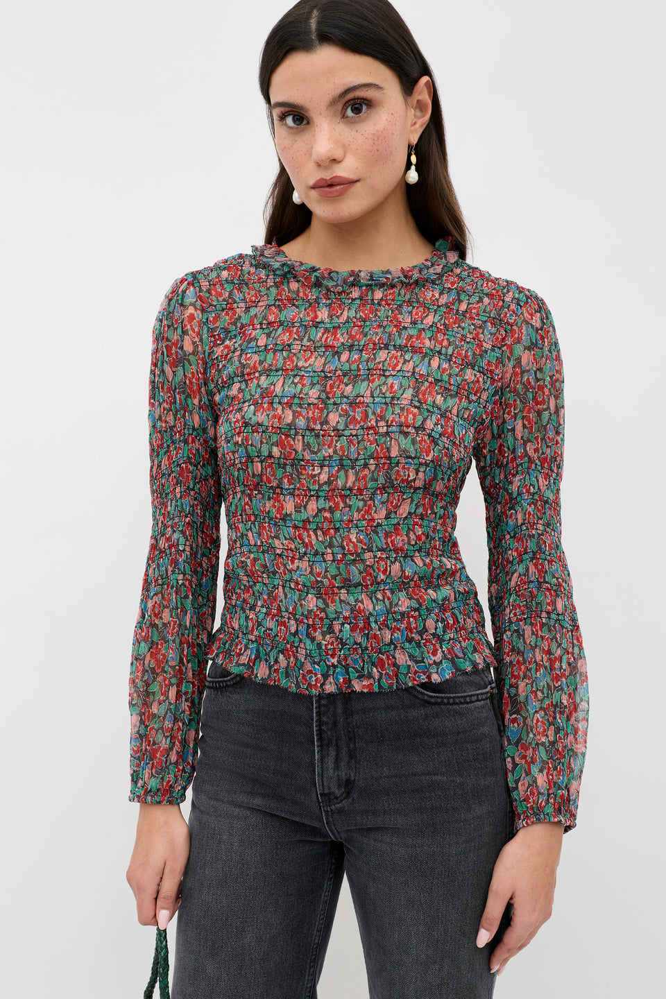 A woman wears the Gardenia - Jeannie Bloom Charcoal sheer, long-sleeve floral blouse with black jeans and drop earrings, standing against a white background and looking at the camera with a neutral expression.