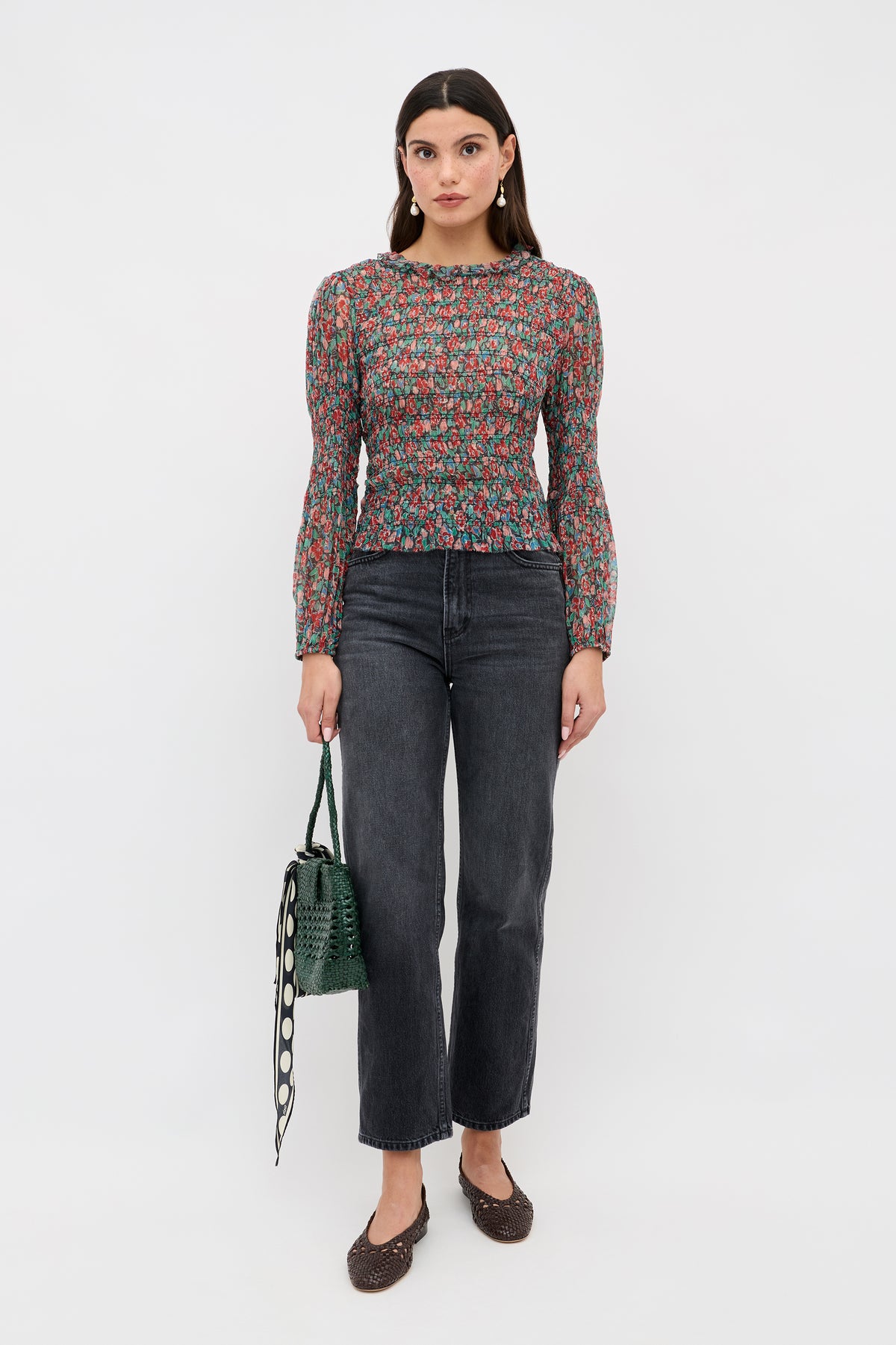 A woman stands against a plain background, wearing the Gardenia - Jeannie Bloom Charcoal blouse, black straight-leg jeans, brown flats, and holding a green textured handbag with a patterned strap.