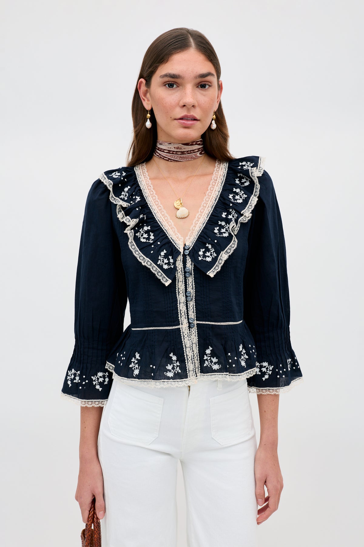 A woman wears the Solene - Flower Trail Embroidery Navy blouse with white pants, accessorized with pearl earrings, layered necklaces, and a brown bag. She has straight brown hair.