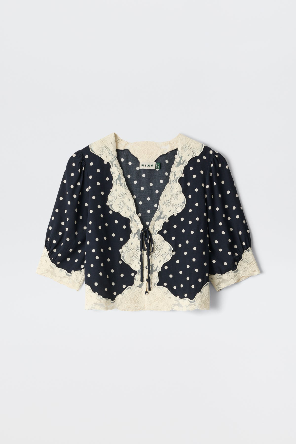 The Ursula - Layla Dot Navy is a navy blouse with white polka dots, puffed half sleeves, cream lace trim on the collar, front, and hem, plus a front tie closure.