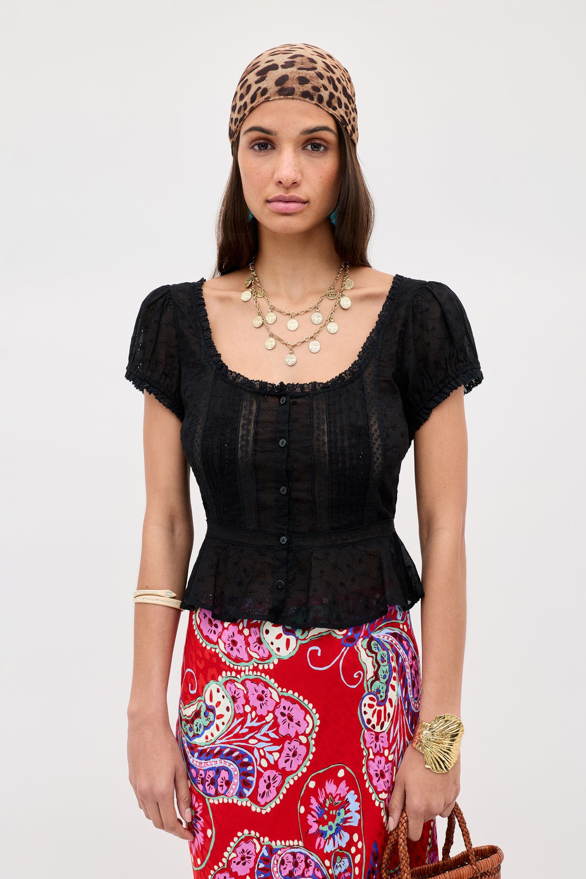 A woman in the Sandi - Black broderie anglaise cotton short-sleeve blouse, a leopard print headscarf, gold layered necklaces, and a red paisley skirt stands against a plain background.