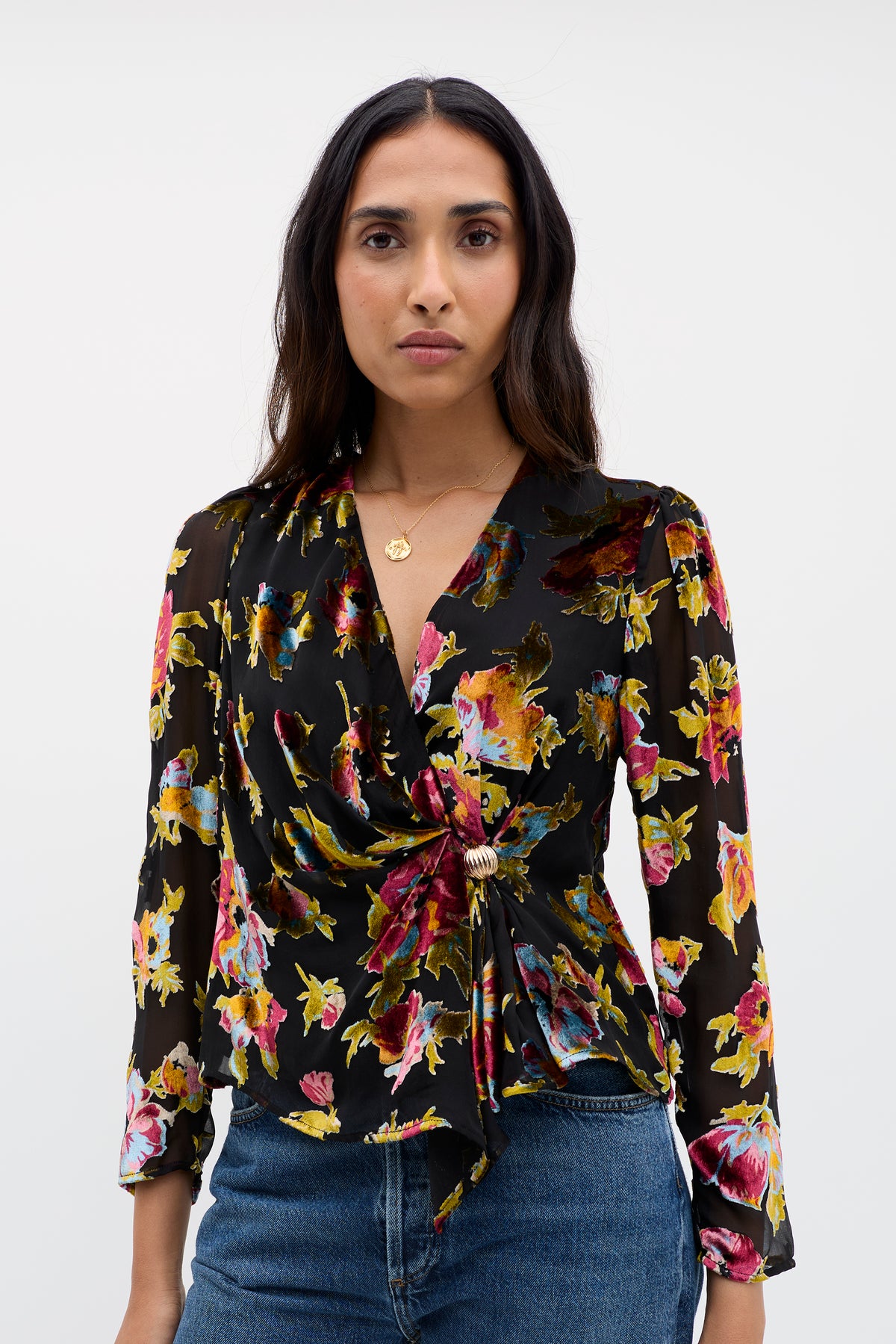 A woman with long dark hair wears the Everlyn - Countryside Devoré Black, a vintage-inspired floral blouse with a side knot detail, paired with blue jeans, standing against a plain white background.