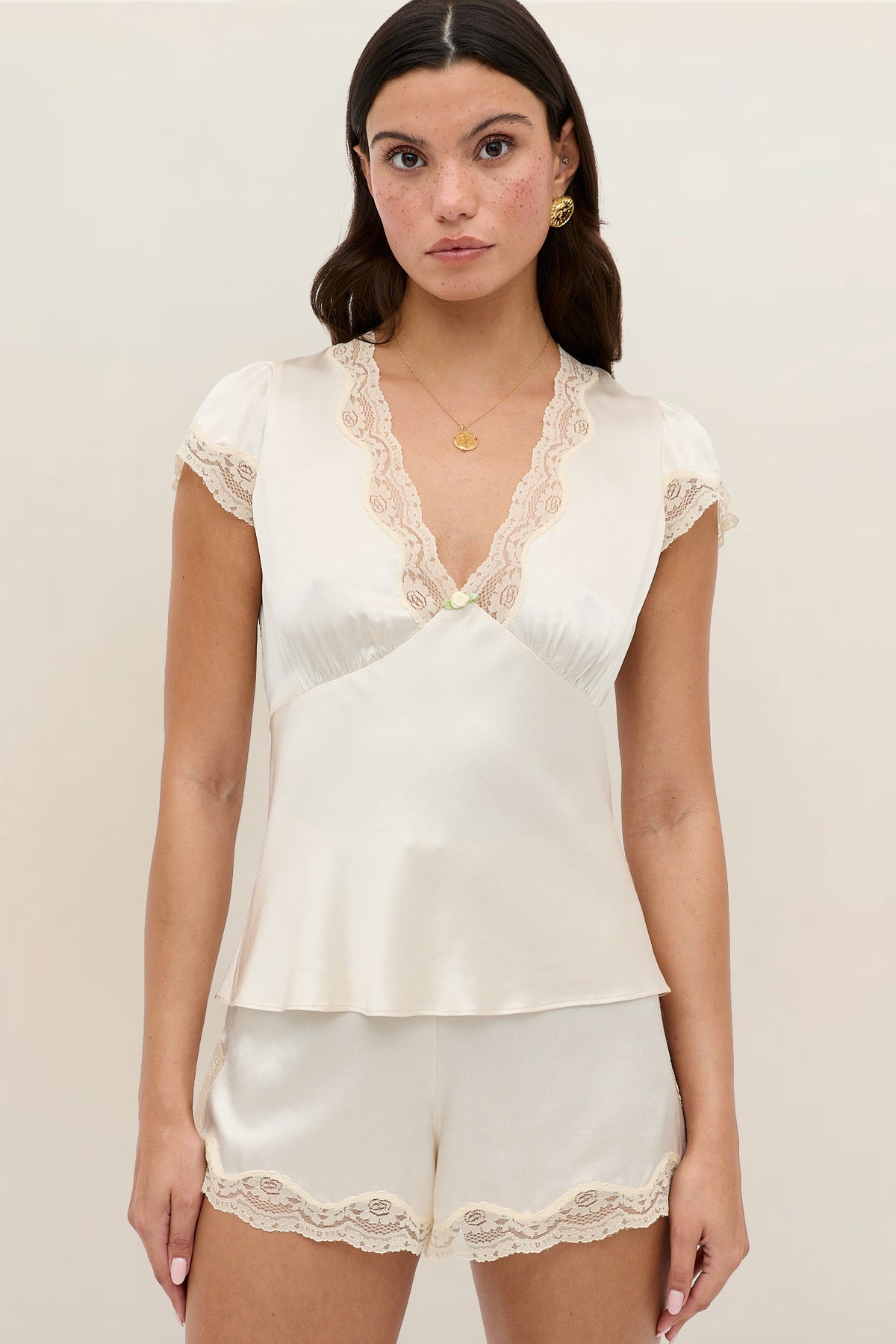 A woman with long dark hair stands against a plain light background, wearing the Karia - Champagne cream satin short-sleeve pajama set with lace trim at the neckline, sleeves, hem, and shorts.