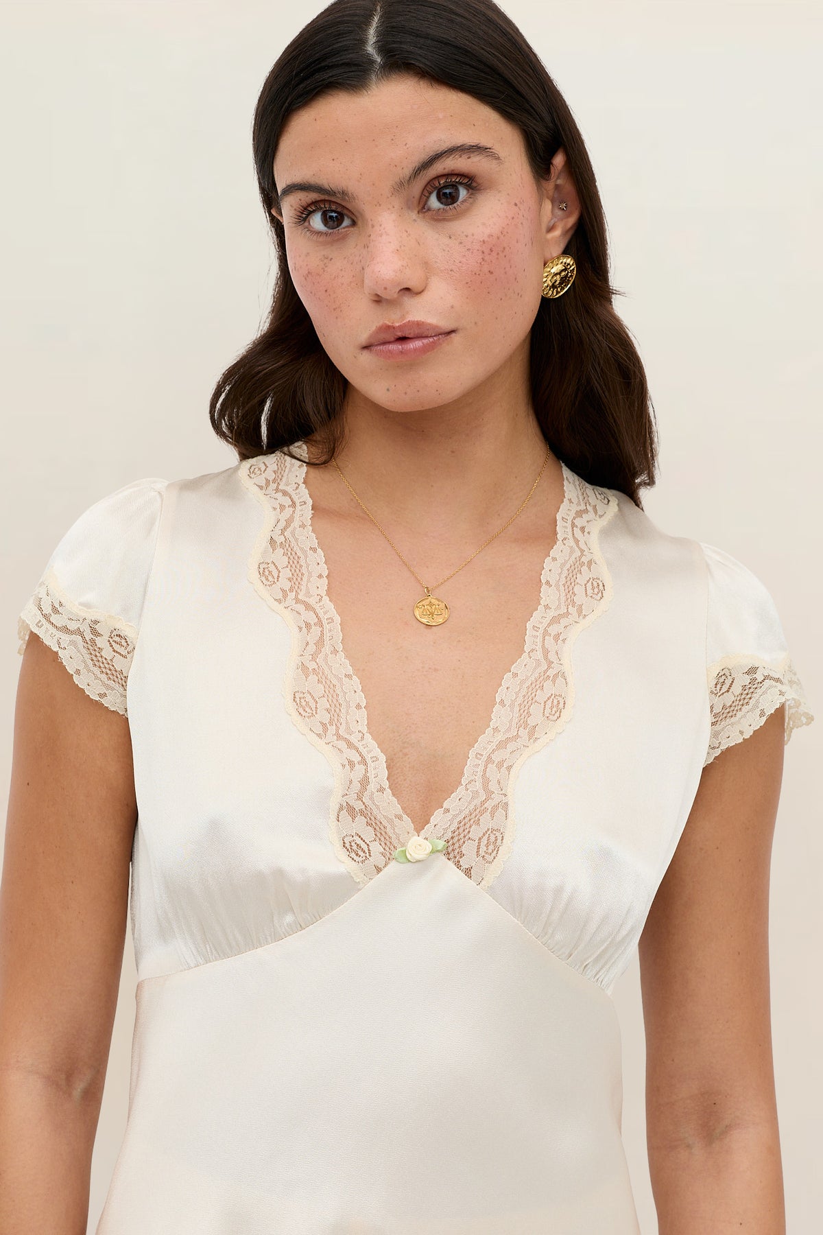 A woman with long brown hair and freckles wears the Karia – Champagne, a cream satin top with lace trim at the neckline and sleeves. She accessorizes with gold earrings and a pendant necklace, standing before a light background.