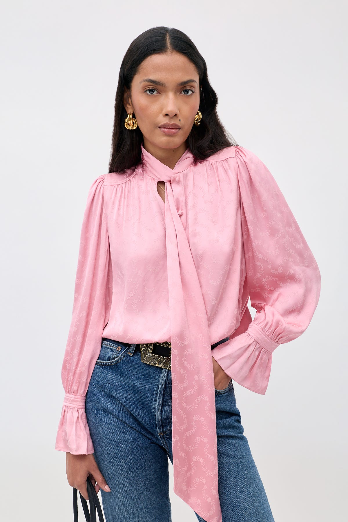Wearing the Evanora - Floral Trail, a silky pink blouse with a necktie detail, paired with blue jeans, gold earrings, and holding a black handbag, a woman stands against a plain light background.