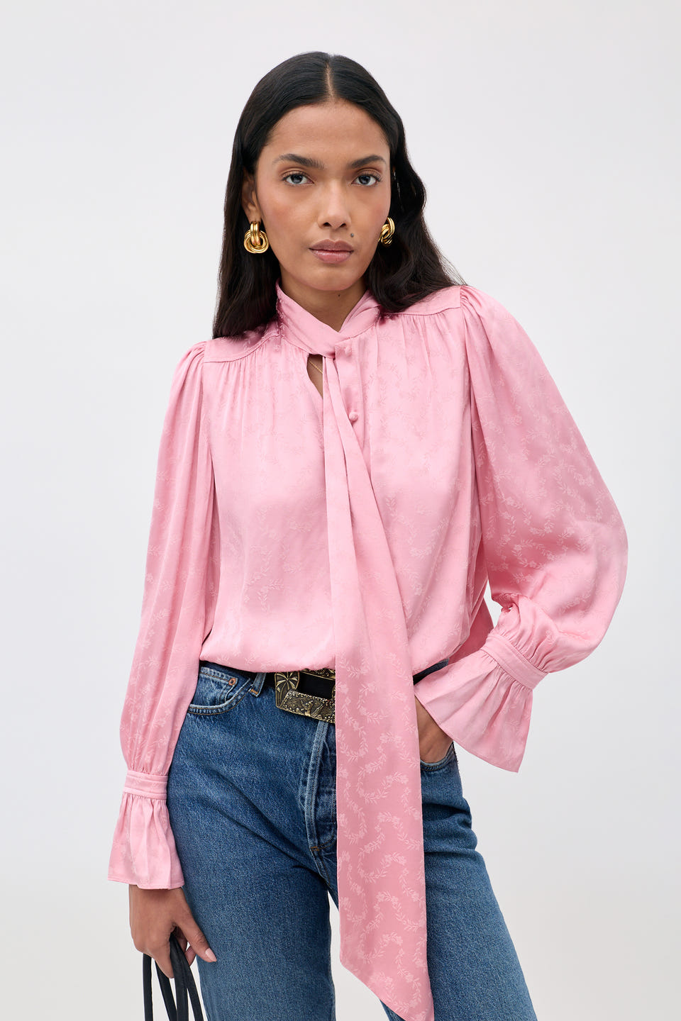 Wearing the Evanora - Floral Trail, a silky pink blouse with a necktie detail, paired with blue jeans, gold earrings, and holding a black handbag, a woman stands against a plain light background.
