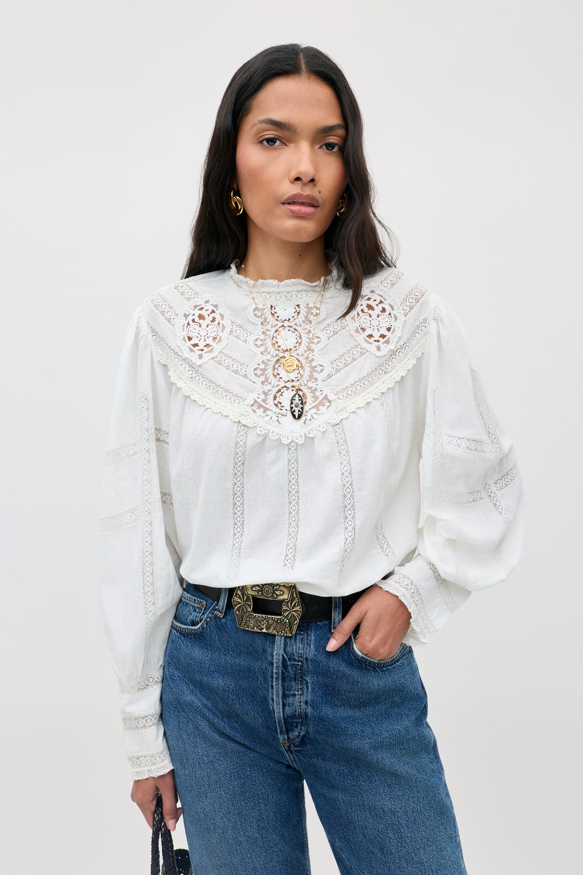 Wearing the Tamzin - Cream blouse, a woman with long dark hair stands against a light gray background, styled with blue jeans, an ornate belt, and gold earrings.