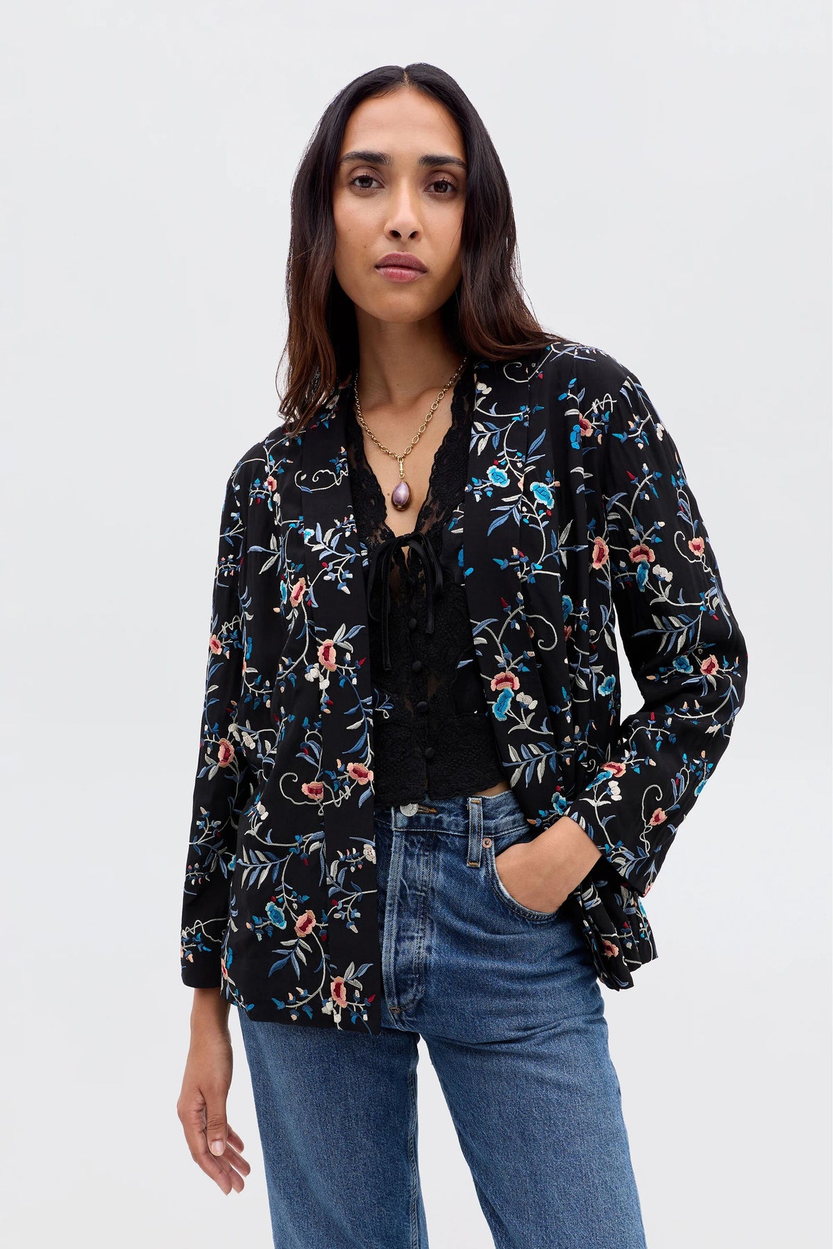 A woman wears the Malachy - Botanical Embroidery Black jacket over a black lace top and blue jeans, standing with one hand in her pocket and confidently facing the camera against a plain light background.