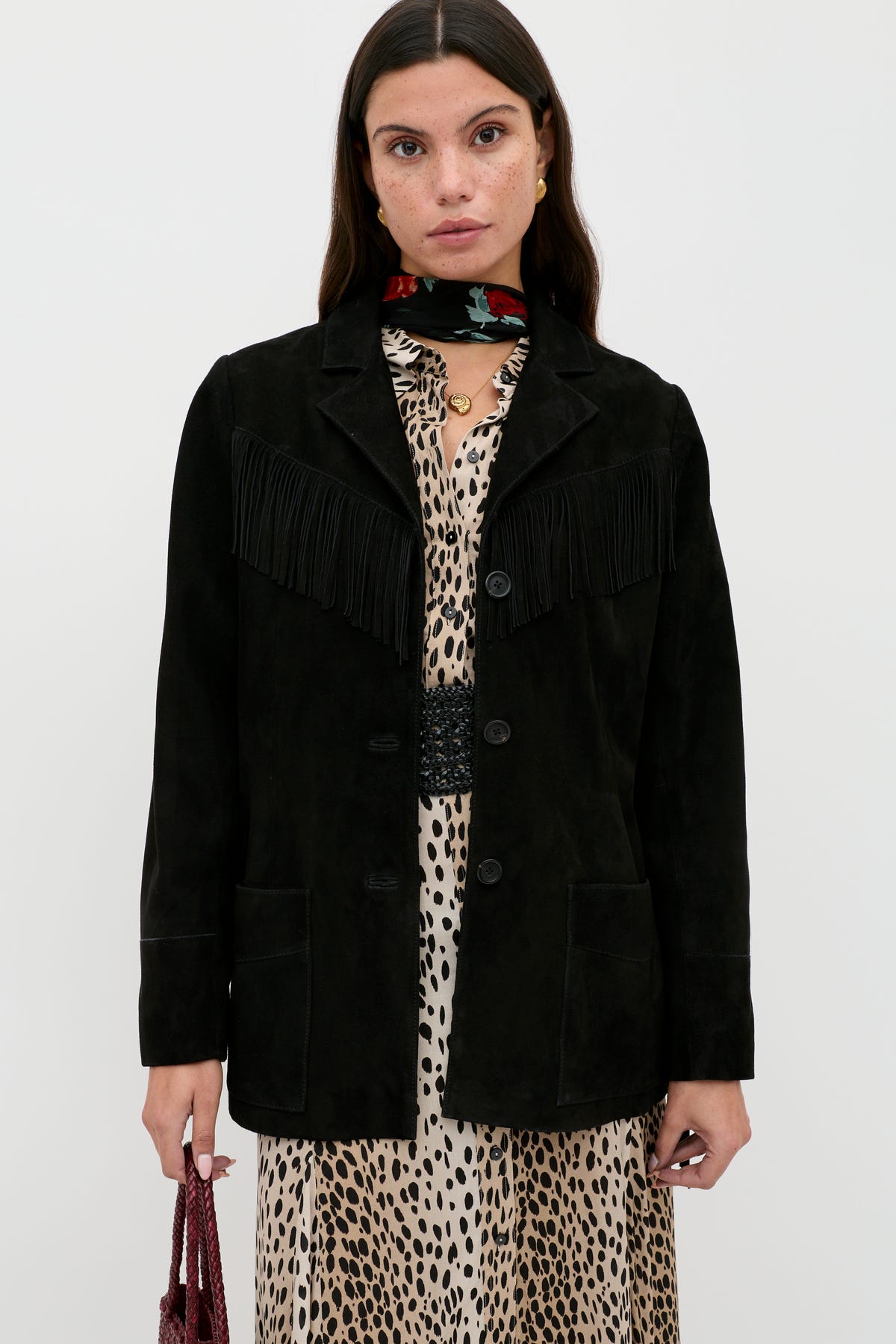 A woman stands against a plain background wearing the Teagan - Black suede jacket with fringe detailing over a leopard print dress, accessorized with a red floral scarf and holding a small textured bag.