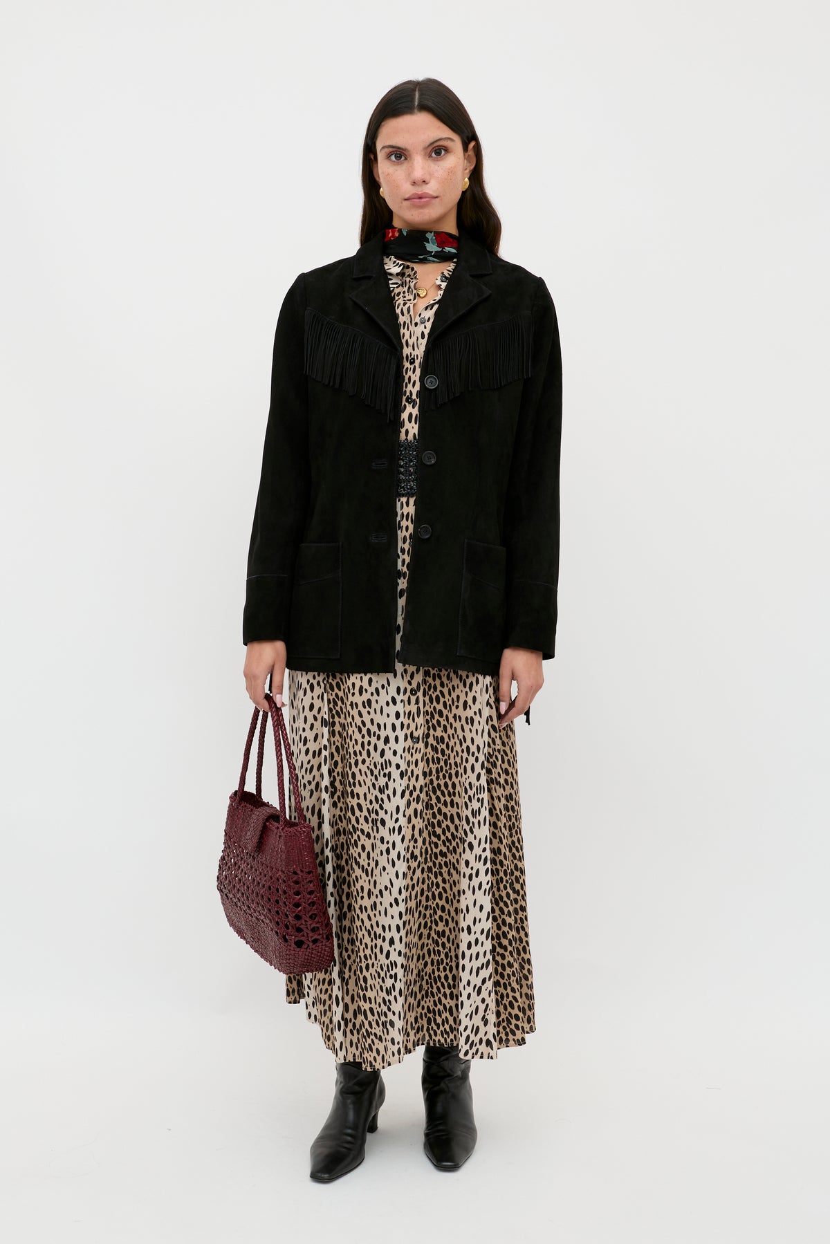 A woman faces forward wearing the Teagan - Black fringed jacket over a long leopard print dress, paired with black ankle boots and a burgundy handbag, against a plain white background.