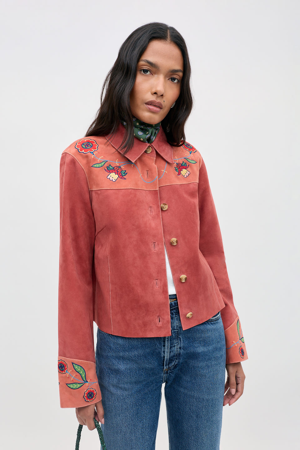 A woman wearing the Karmen - Folk Bloom Appliqué Brown jacket, featuring floral embroidery on the shoulders and sleeves, paired with blue jeans and a green scarf, stands against a plain light background.