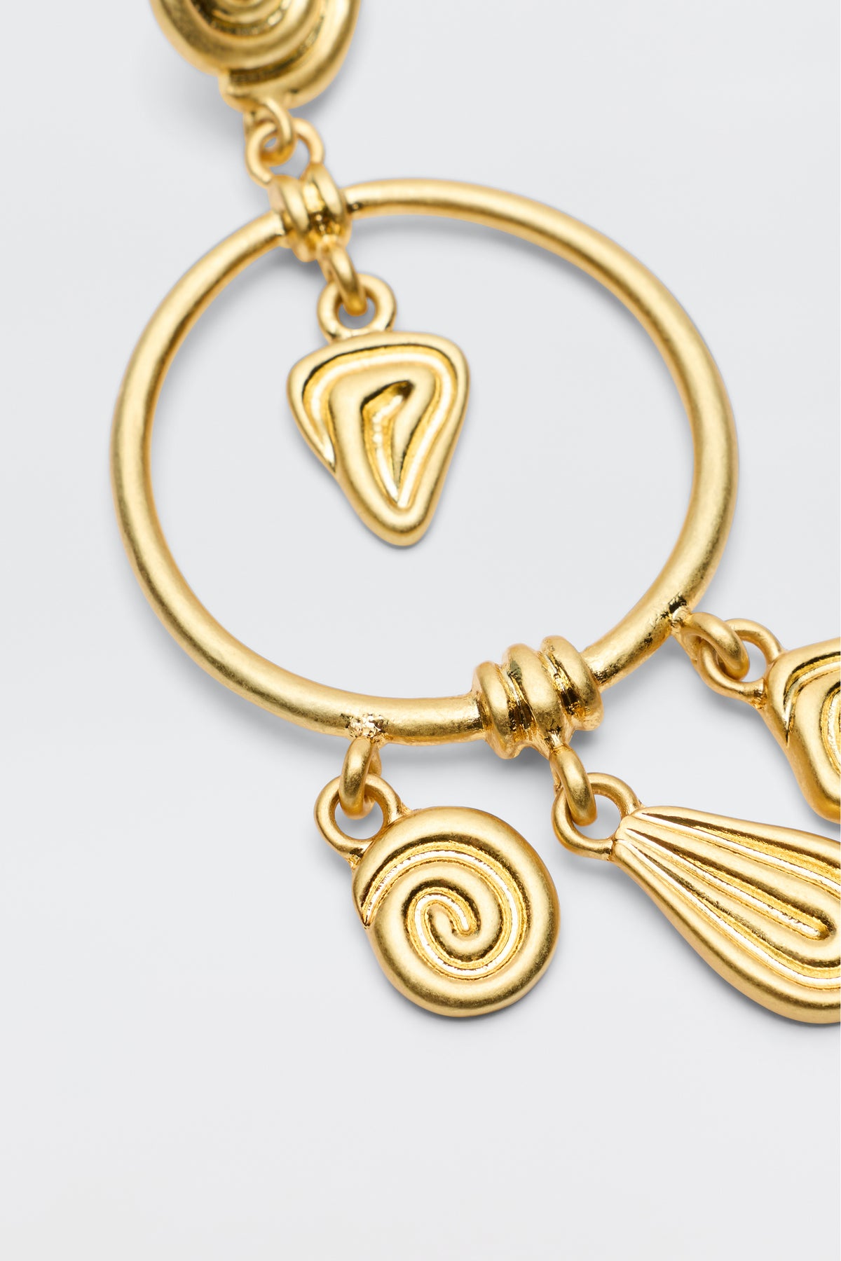 A close-up of the Thome - Gold earring by RIXO, showcasing a large hoop with three dangling abstract charms—a swirl, teardrop, and irregular oval—creating a striking bohemian look against a light background.