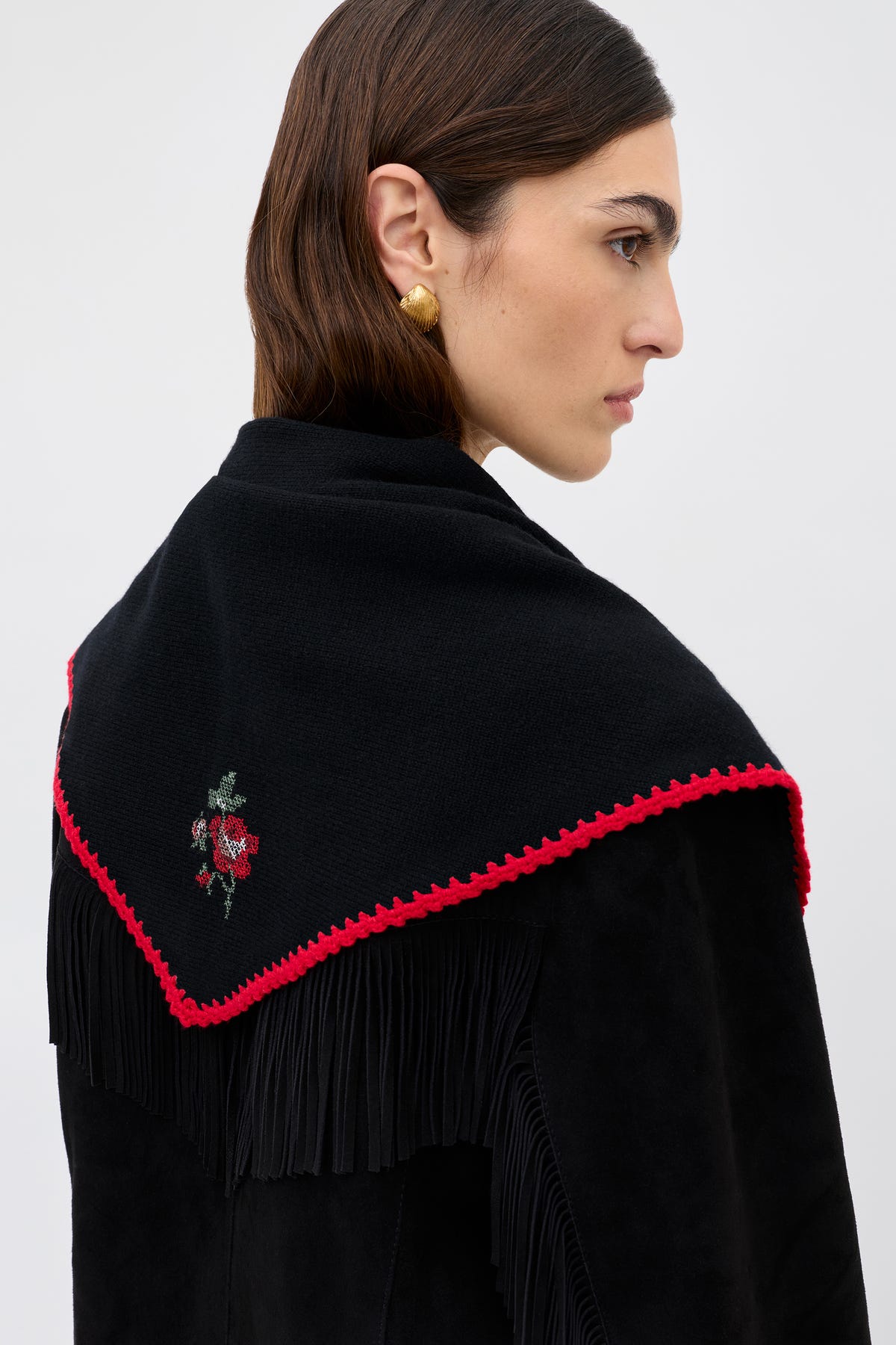 Wearing the Lianora Abi Rose Knit Black—a black shawl with red trim, fringe, and a small embroidered rose on the back—a person with straight brown hair completes their look with a heart-shaped gold earring.