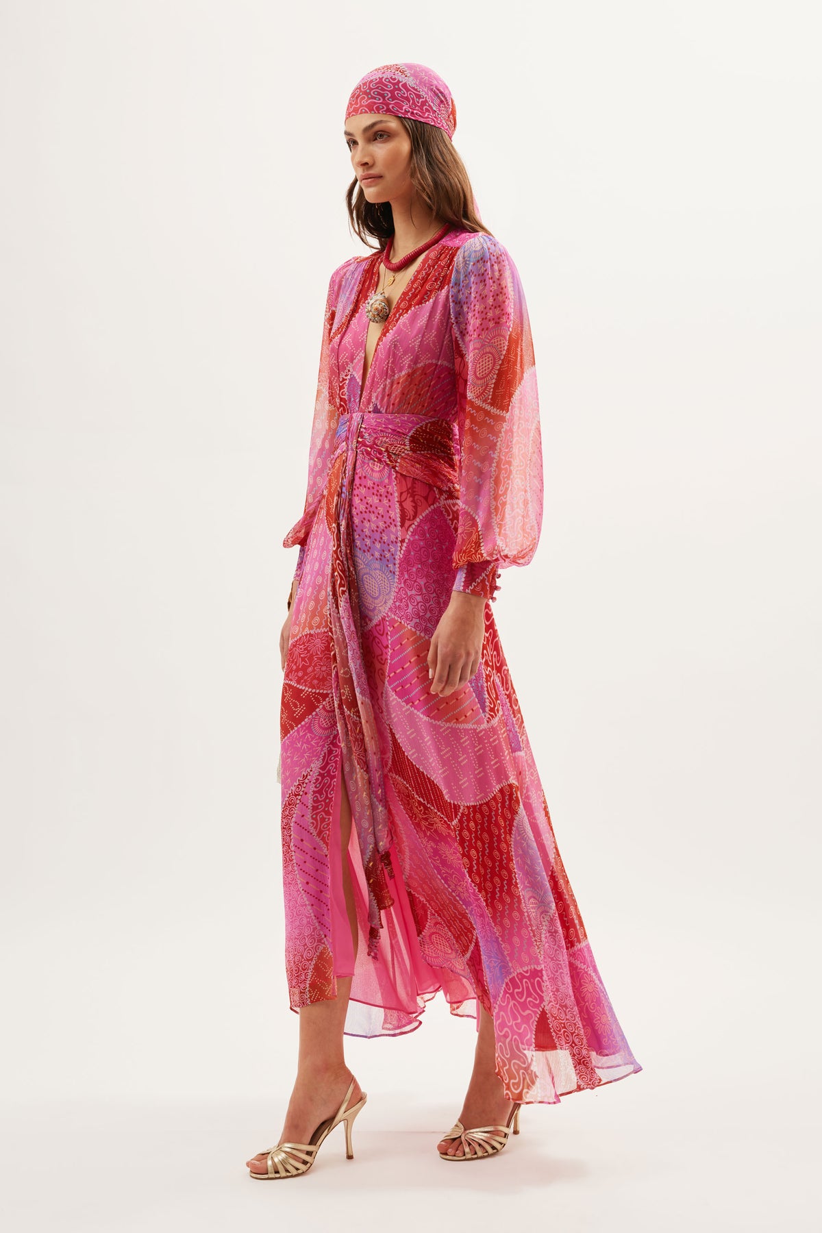 A woman in the Meera - Ibiza Night Sunset gown, crafted from crinkled chiffon with long sleeves and a matching headscarf, stands in heeled sandals against a plain white background.