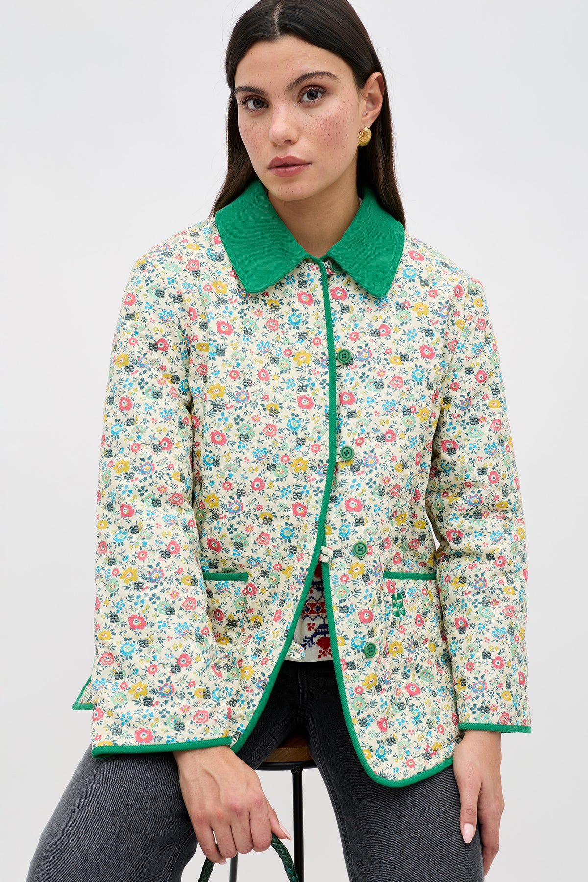A woman with long dark hair sits on a stool wearing the Earla - Amalfi Floral Cream jacket, featuring a bright green collar and trim, paired with dark pants, against a plain light background.