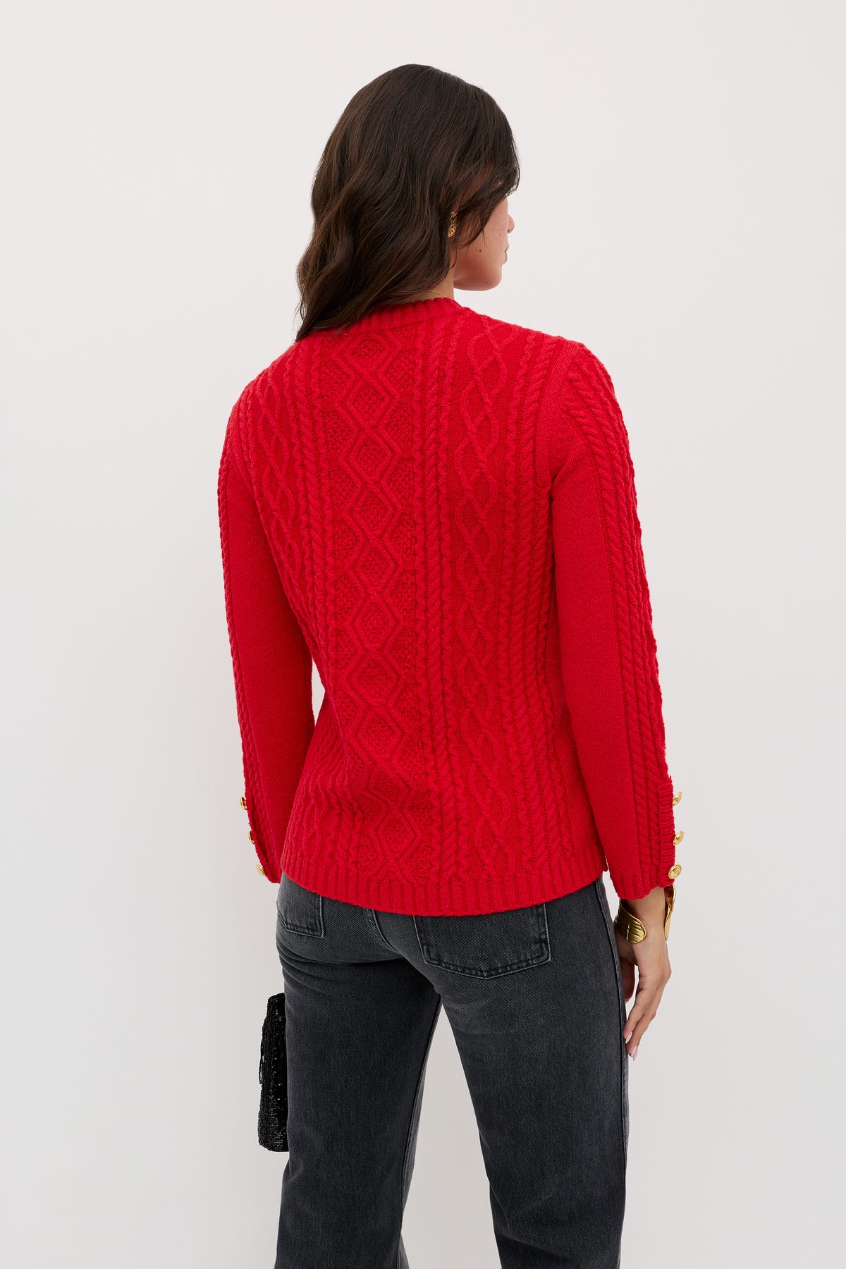 A woman with wavy brown hair stands with her back to the camera, wearing the Maurya - Red cable-knit sweater and black jeans. She holds a small black bag in her left hand against a plain white background.