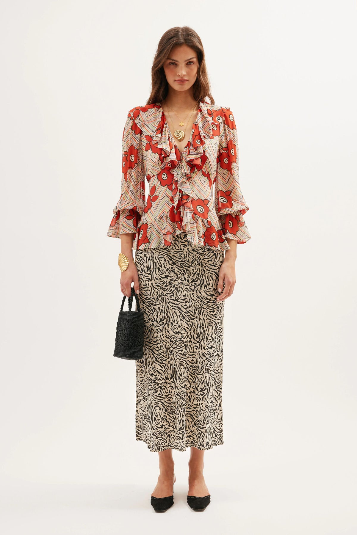 A woman stands against a white background wearing a red floral ruffled blouse, the Ardith - Sun Leo Tan skirt with an elasticated waistband, black pointed shoes, gold jewelry, and holding a small black bag.