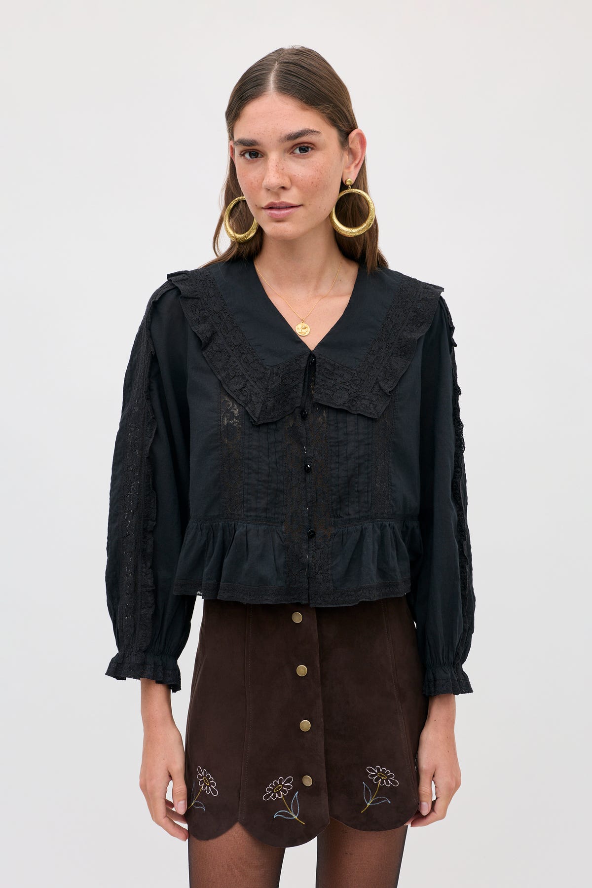 A woman wears a Caris - Shasta Daisy Embroidery Chocolate skirt with small floral embroidery along the scalloped hem, paired with a black long-sleeved blouse featuring a lace-trimmed collar.