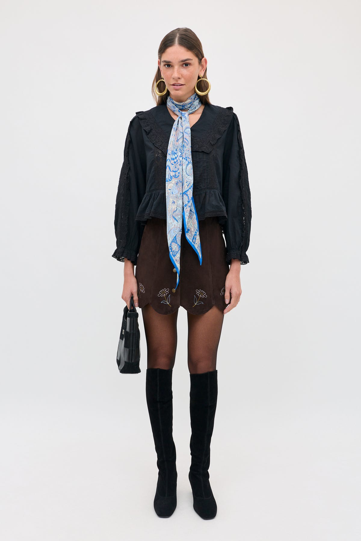 A model wears a black ruffled long-sleeved top, a Caris - Shasta Daisy Embroidery Chocolate skirt with a scalloped hem, a patterned blue neck scarf, and knee-high black boots while holding a small black handbag.