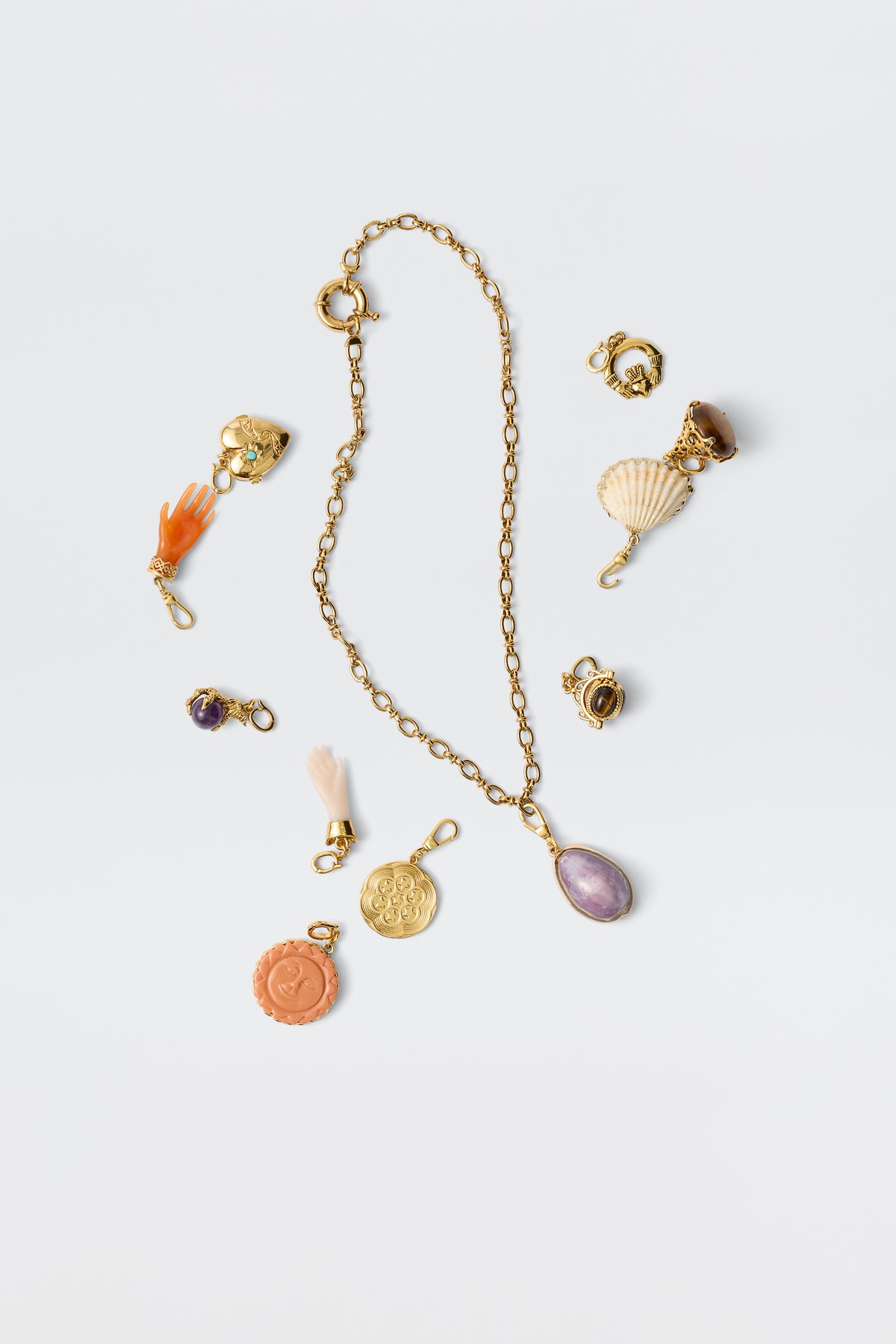 A gold chain necklace with an orange oval pendant is surrounded by vintage charms—a seashell, coin, hand, and tassel—from the Lillias - Orange collection, displayed on a plain white background.