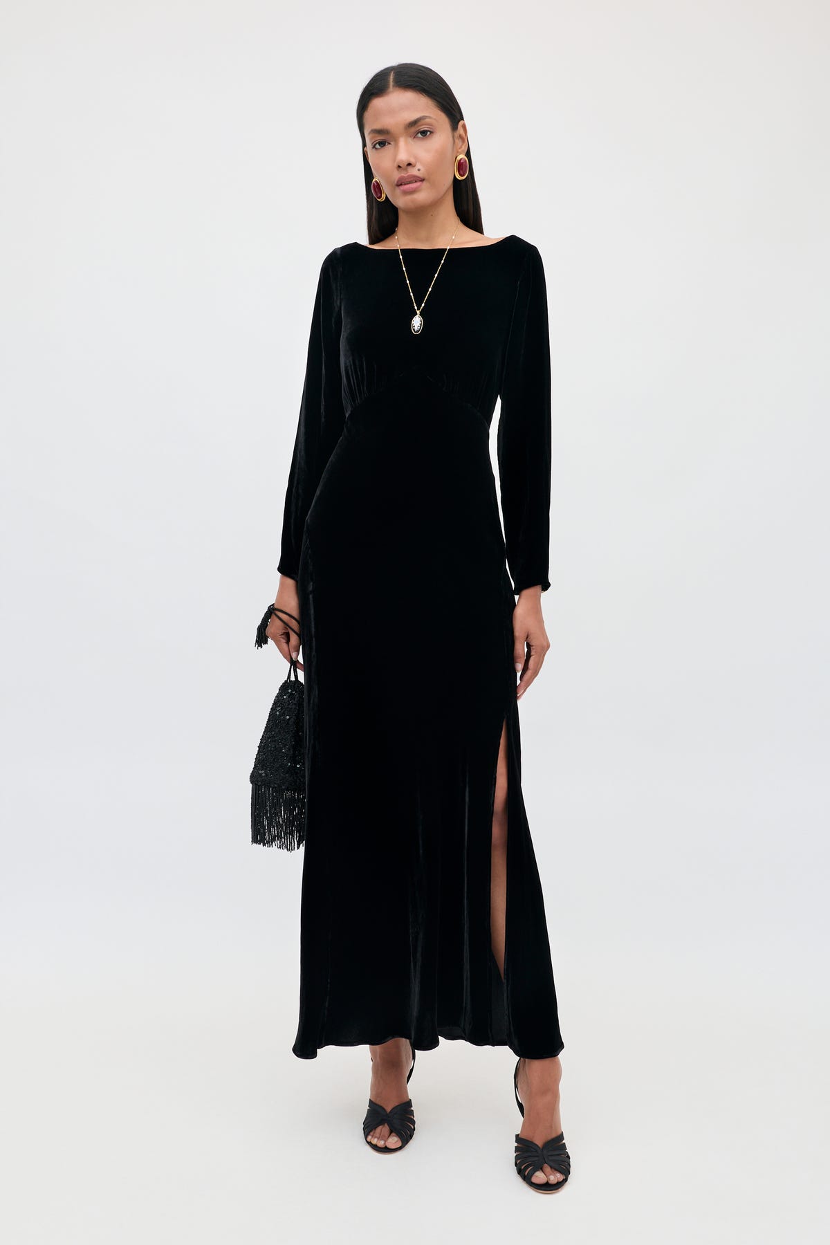 Wearing the Devina - Black gown, a woman stands against a plain background. The long velvet dress features long sleeves and a side slit, highlighting her sophisticated style. She completes the look with black heels, a black handbag, gold hoop earrings, and a pendant necklace.