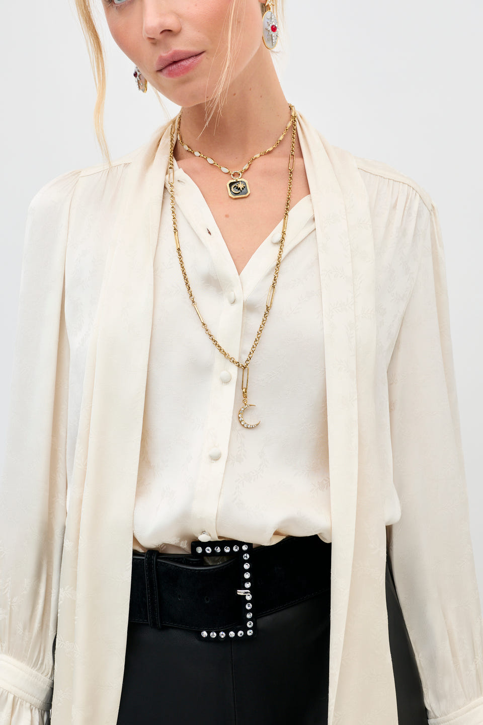 A woman wears the Eta - Gold necklace layered over a cream blouse, paired with statement earrings and a black belt with silver studs. Her face is partially visible, and her blonde hair is styled in a loose, casual look.