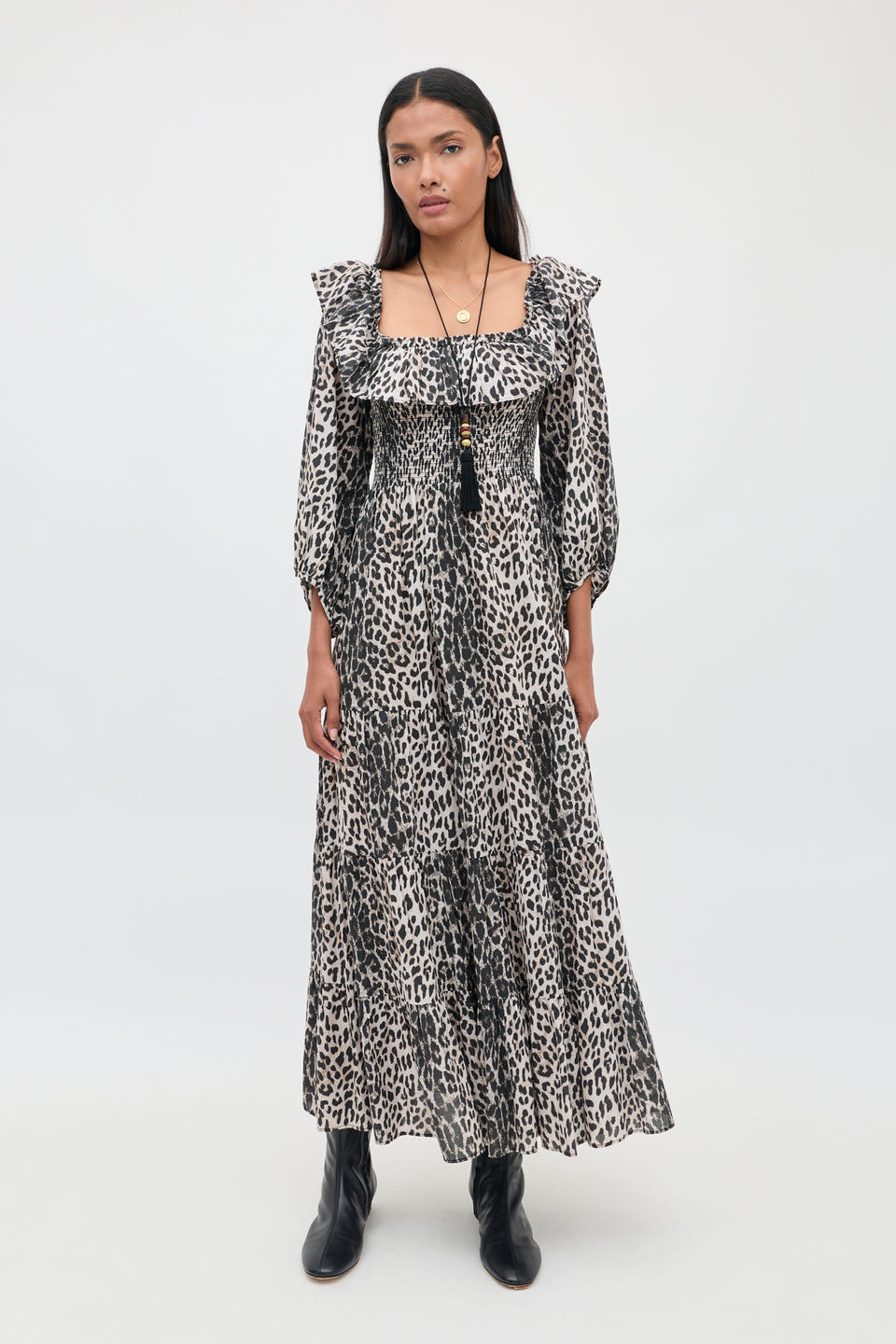 A person stands against a plain background wearing the Gabriel - Nina Leopard Mocha dress with ruffled sleeves and black ankle boots. They have long dark hair and are accessorized with layered necklaces.