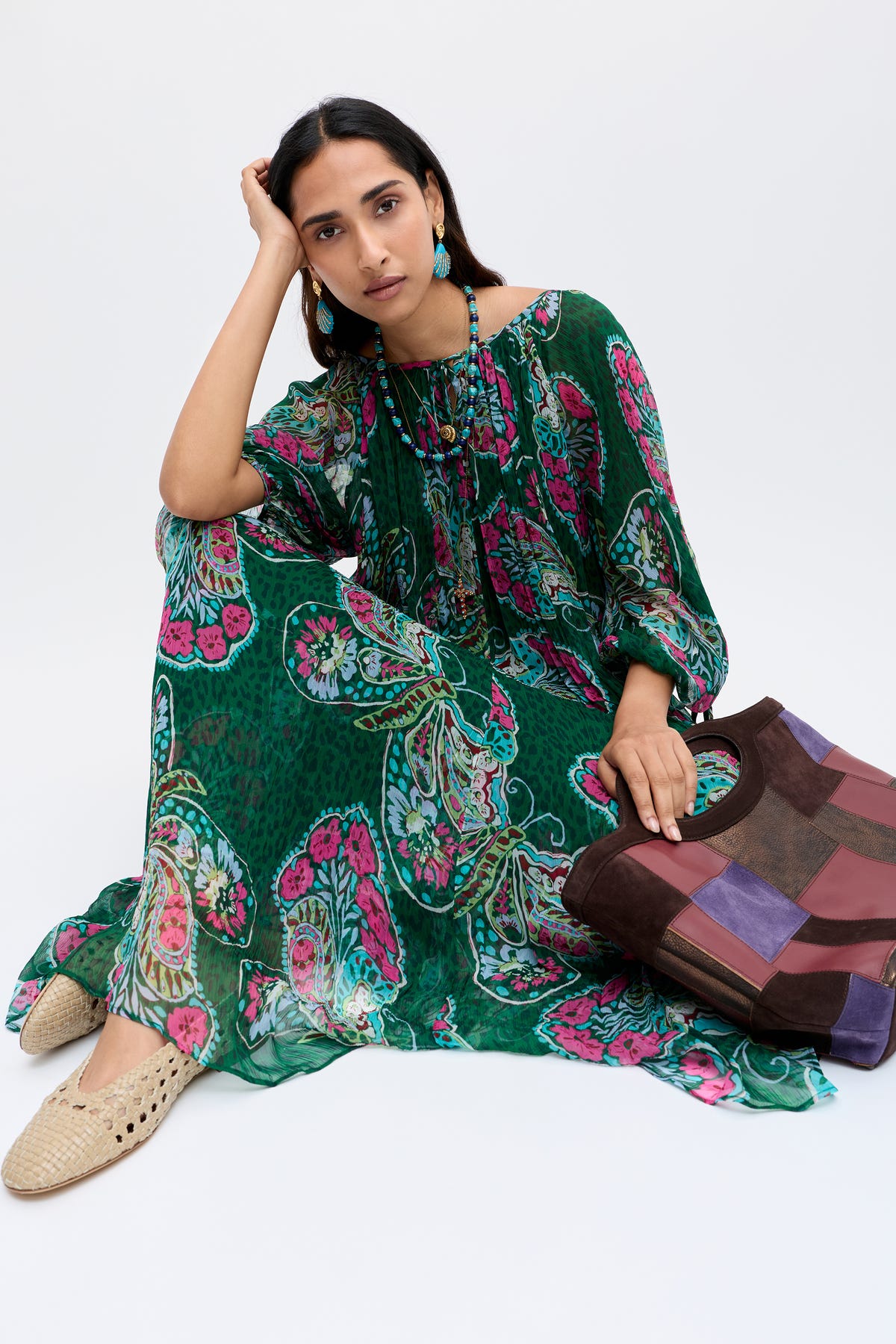 A woman in the Hayden - Butterfly Bloom Green dress sits on the floor, leaning her head on her hand. She accessorizes with woven shoes, blue earrings, colorful beaded necklaces, and a large patchwork bag against a white background.