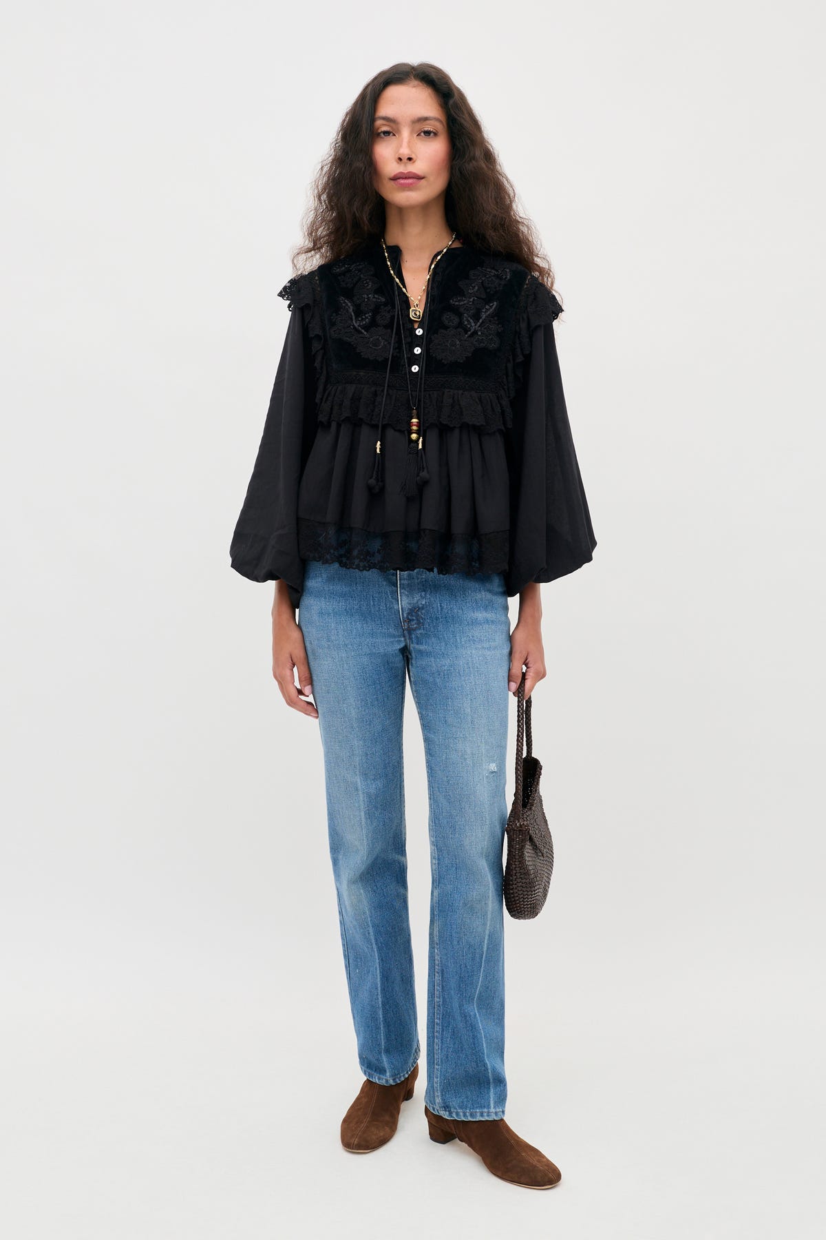 A person with long wavy hair wears the Diann - Lilly Embroidery Black blouse, paired with blue jeans and brown ankle boots, holding a small dark handbag while standing against a plain light background.