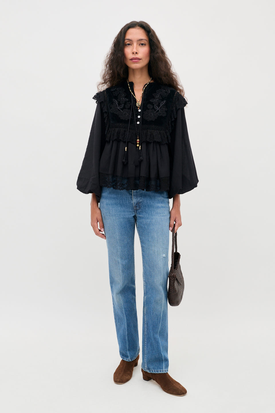 A person with long wavy hair wears the Diann - Lilly Embroidery Black blouse, paired with blue jeans and brown ankle boots, holding a small dark handbag while standing against a plain light background.