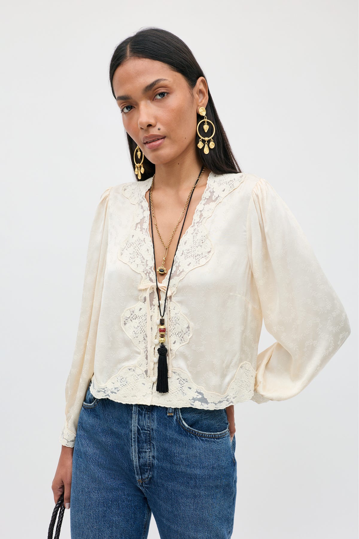A woman wearing the Tove - Floral Trail Jacquard Champagne blouse with long sleeves, styled with blue jeans, layered necklaces, and large gold earrings, stands against a plain light background.