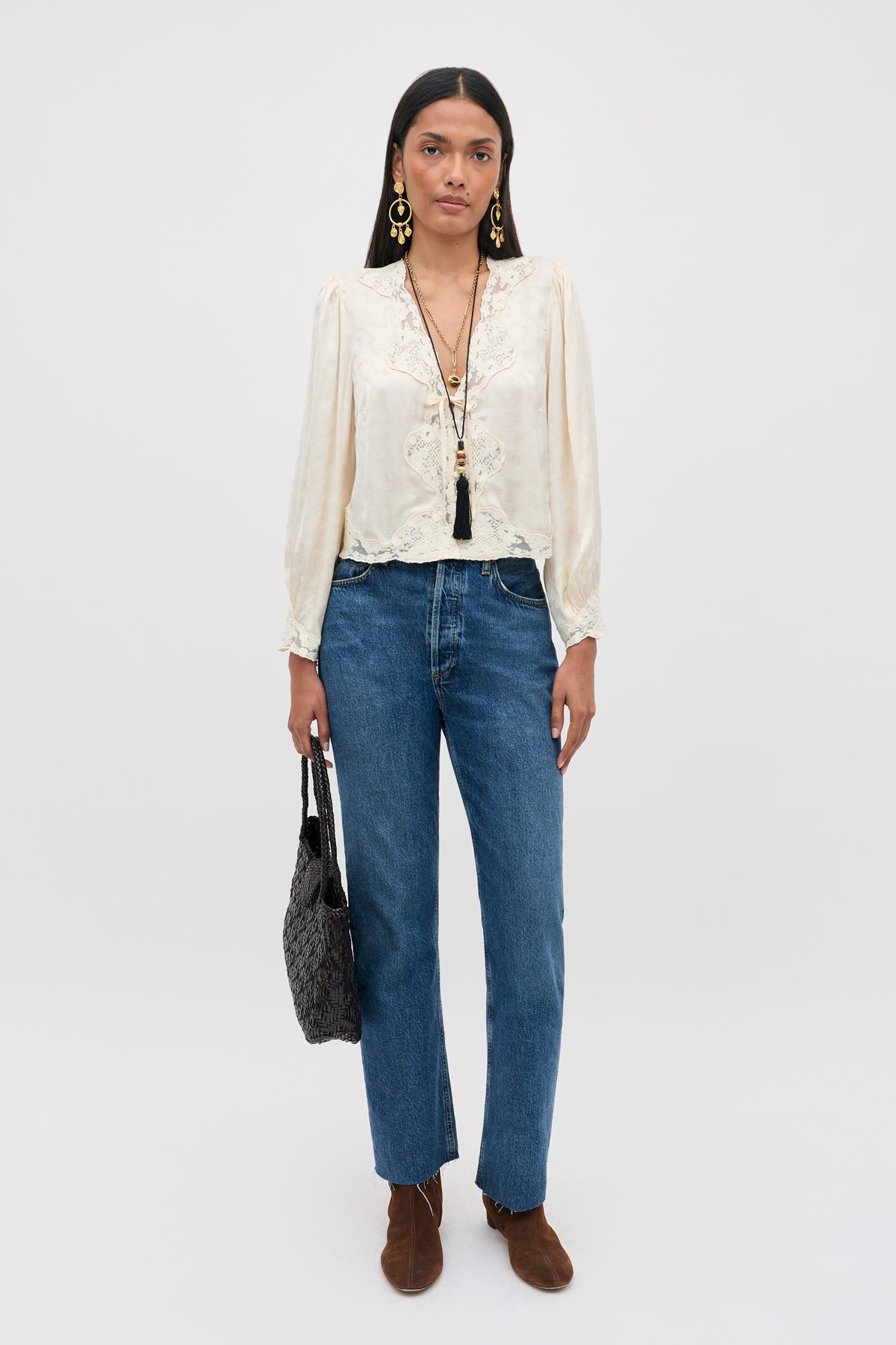 A woman stands against a plain background, wearing the Tove - Floral Trail Jacquard Champagne blouse paired with blue jeans, brown boots, statement earrings, layered necklaces, and holding a textured black bag.