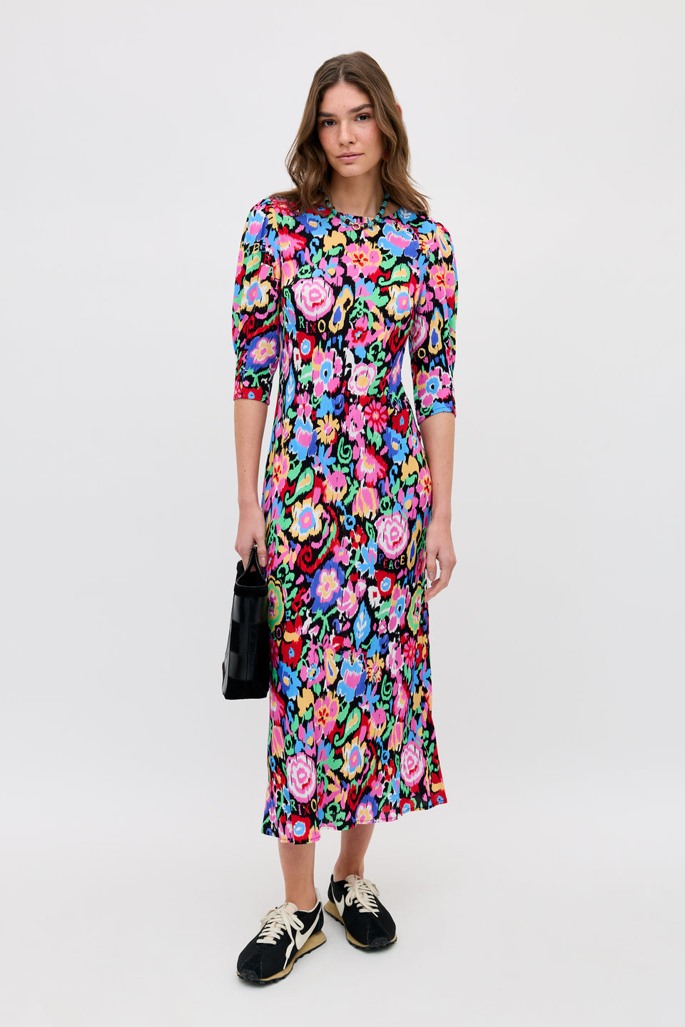A woman stands against a white background in the Jess - Ikat Bloom Charcoal, an ankle-length floral dress with elbow-length sleeves. She accessorizes with a black handbag and black sneakers with white soles.