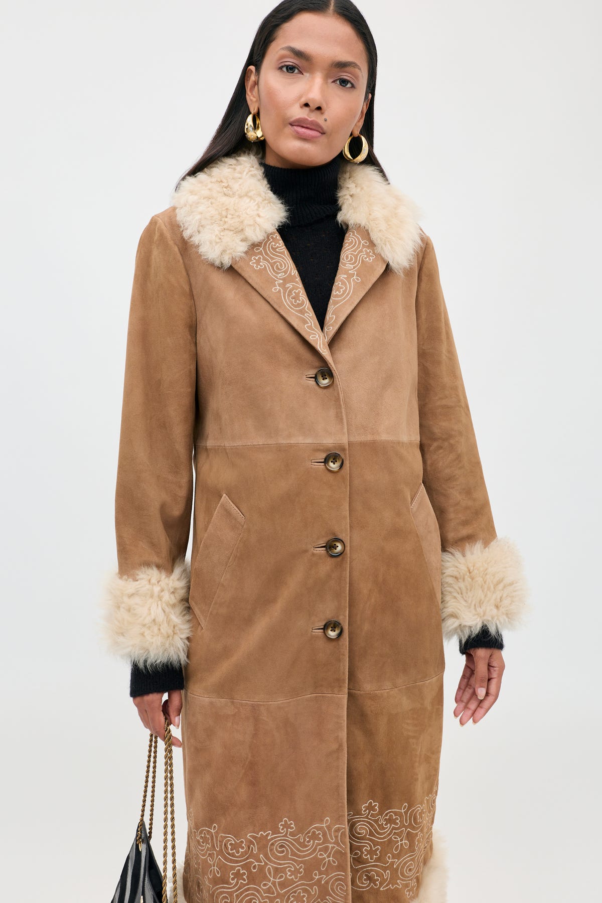 Woman wearing the Cienna - Camel coat, a tan suede piece with a white faux fur collar and cuffs, paired with gold hoop earrings and a black handbag. This chic shearling coat features button closure and decorative embroidery near the hem.