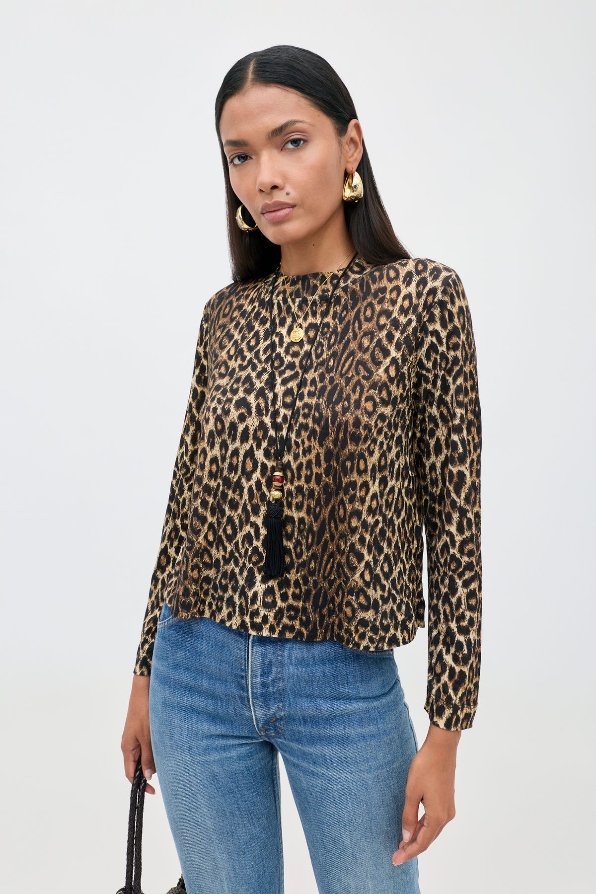 A woman is wearing the Phaedra - Lyra Leopard Black long-sleeve crop top, crafted from soft Tencel Jersey, paired with blue jeans, gold hoop earrings, and a long black tassel necklace as she stands against a plain white background.