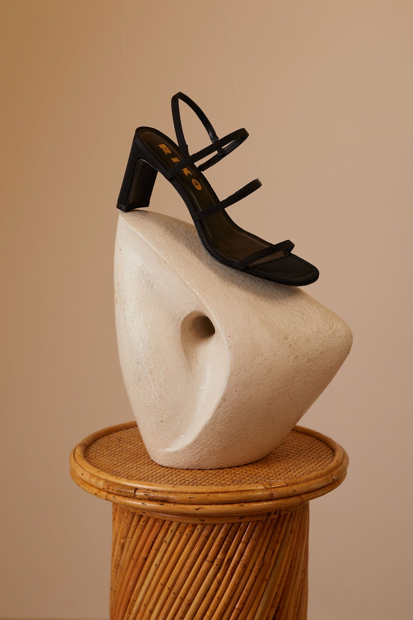 The Ciervo - Black, a vintage-inspired strappy leather sandal, is shown balanced on a smooth abstract beige stone sculpture that rests on a round wicker pedestal against a plain beige background.