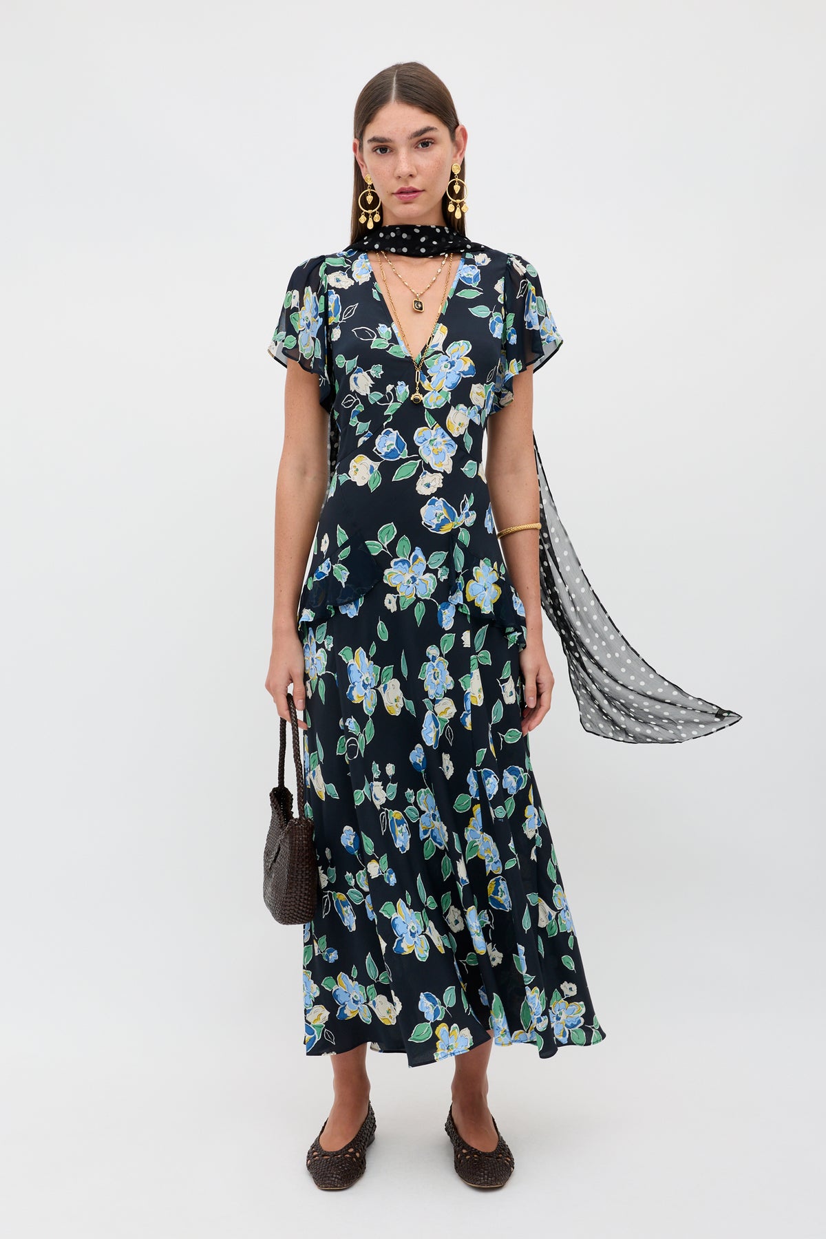 A woman stands against a white background wearing the Evie - Kimmie Floral Navy dress, a sheer black polka dot scarf, gold earrings, black shoes, and holding a brown woven handbag.