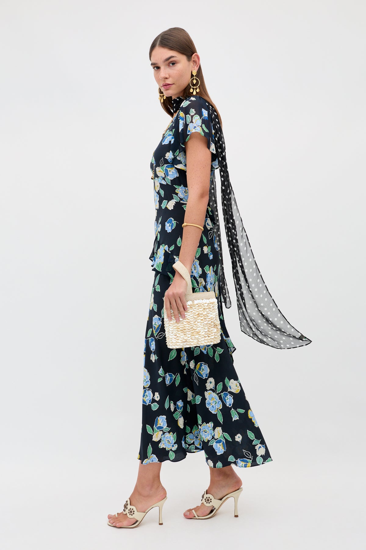 Wearing the Evie - Kimmie Floral Navy dress, a woman in profile pairs it with gold earrings, beige heels, a long polka dot scarf, and holds a textured cream clutch, all set against a plain white background.