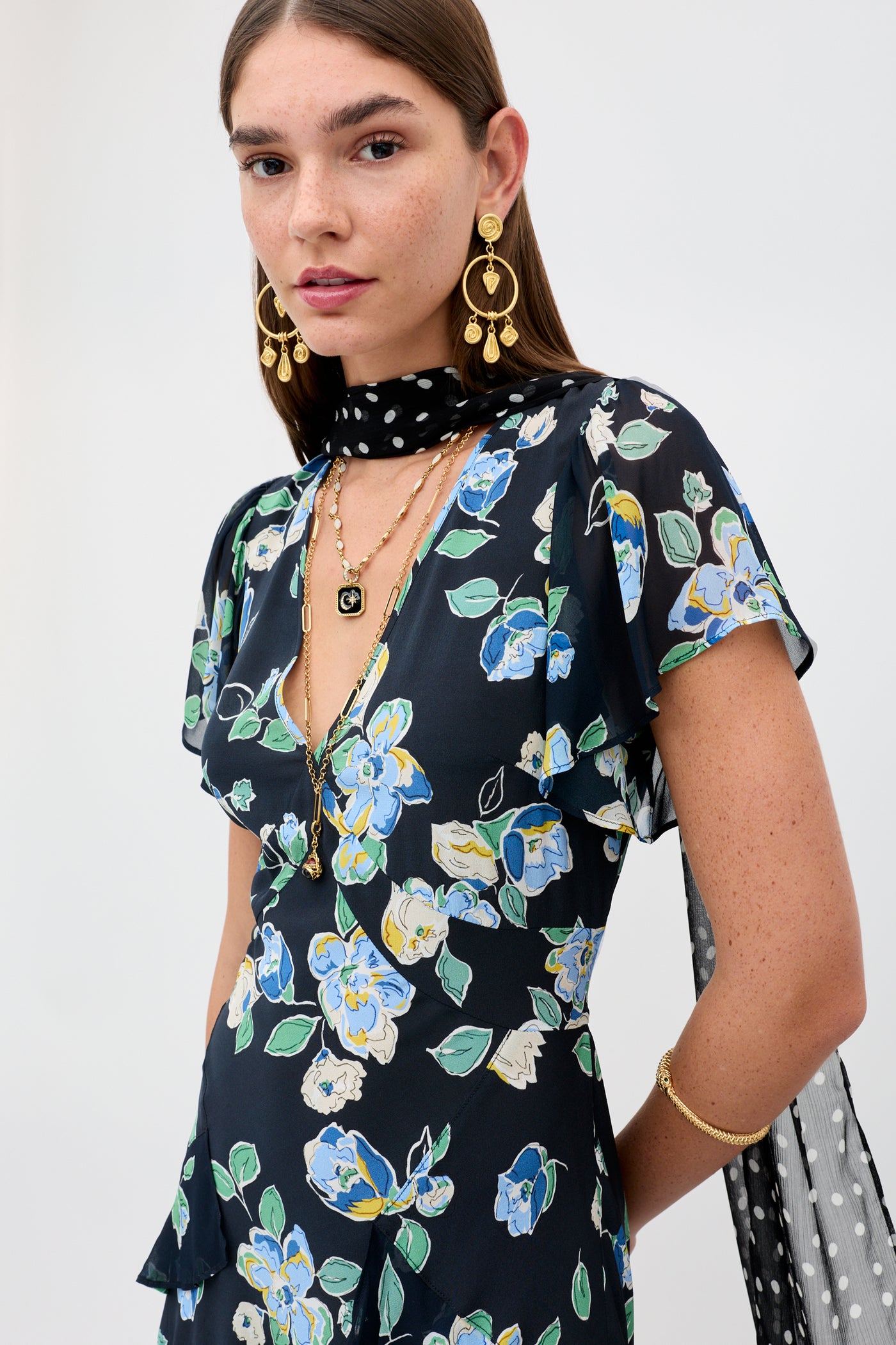 A woman with straight brown hair wears the Evie - Kimmie Floral Navy dress, layered gold jewelry, and a black scarf with white polka dots against a plain light background.