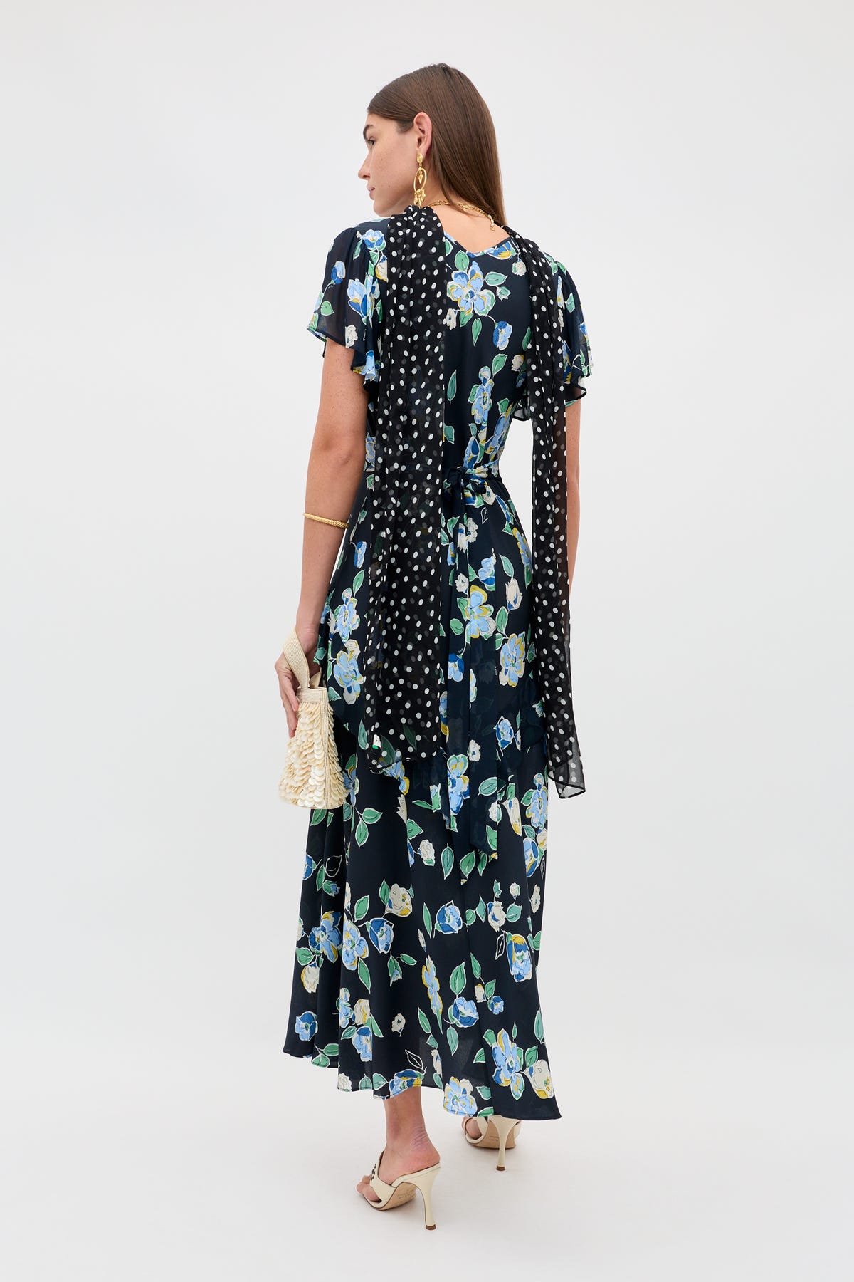 A woman with long brown hair wears the Evie - Kimmie Floral Navy dress, a black scarf with white polka dots, cream heeled sandals, gold jewelry, and carries a woven handbag.