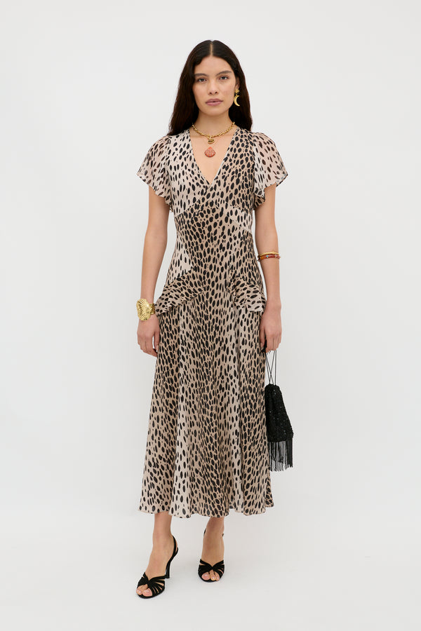 A woman with long, dark hair wears the Evie - Antelope Spot Black dress against a white background, paired with black heeled sandals, gold jewelry, and a black fringed handbag. Her neutral expression completes the effortless bohemian look.