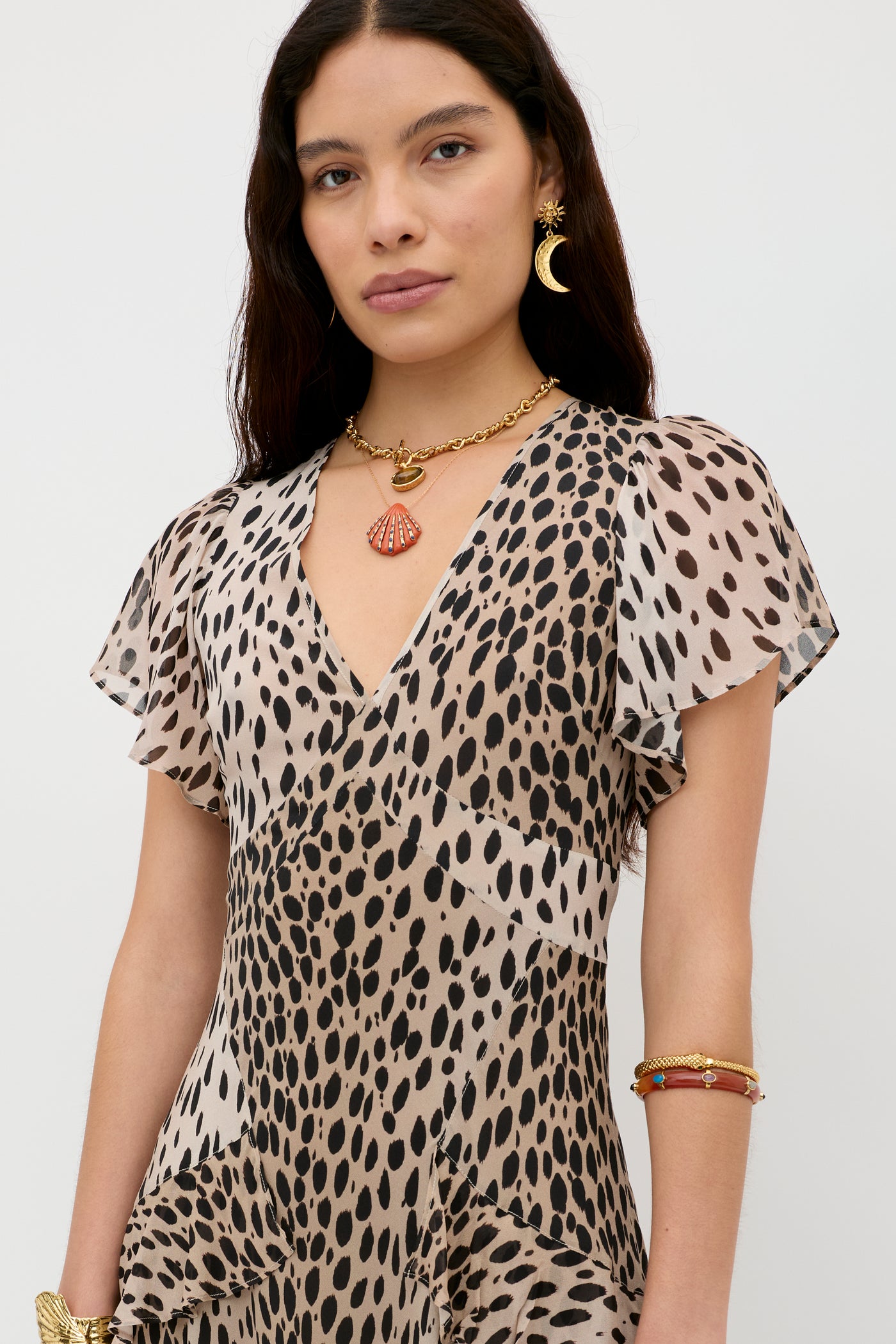 A woman with long dark hair wears the Evie - Antelope Spot Black dress, gold crescent moon earring, shell necklace, and bohemian bracelets, standing against a plain white background.